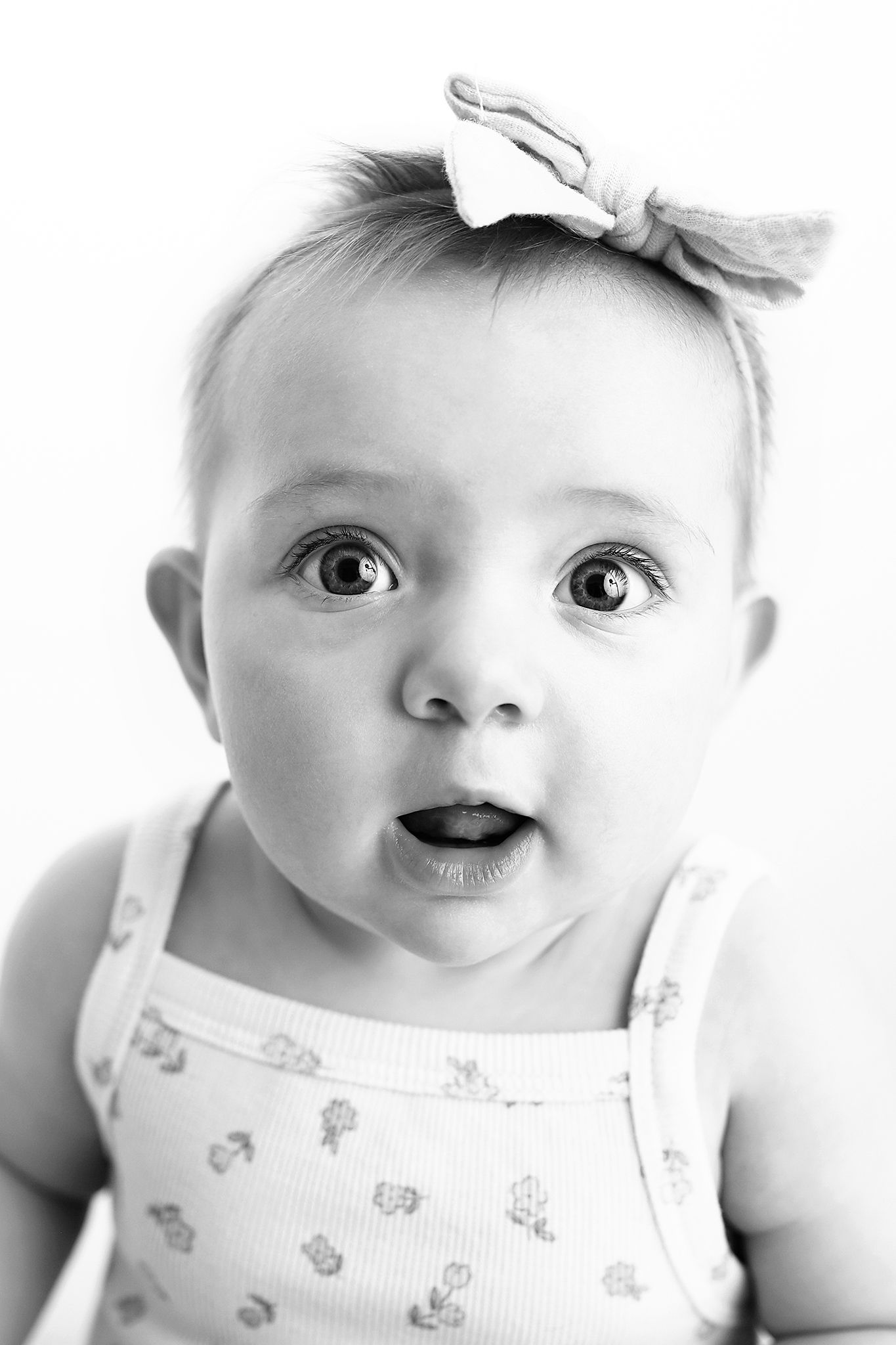 Black and white portrait of a wide-eyed baby with an open mouth, wearing a bow headband and a sleeveless floral-print top.