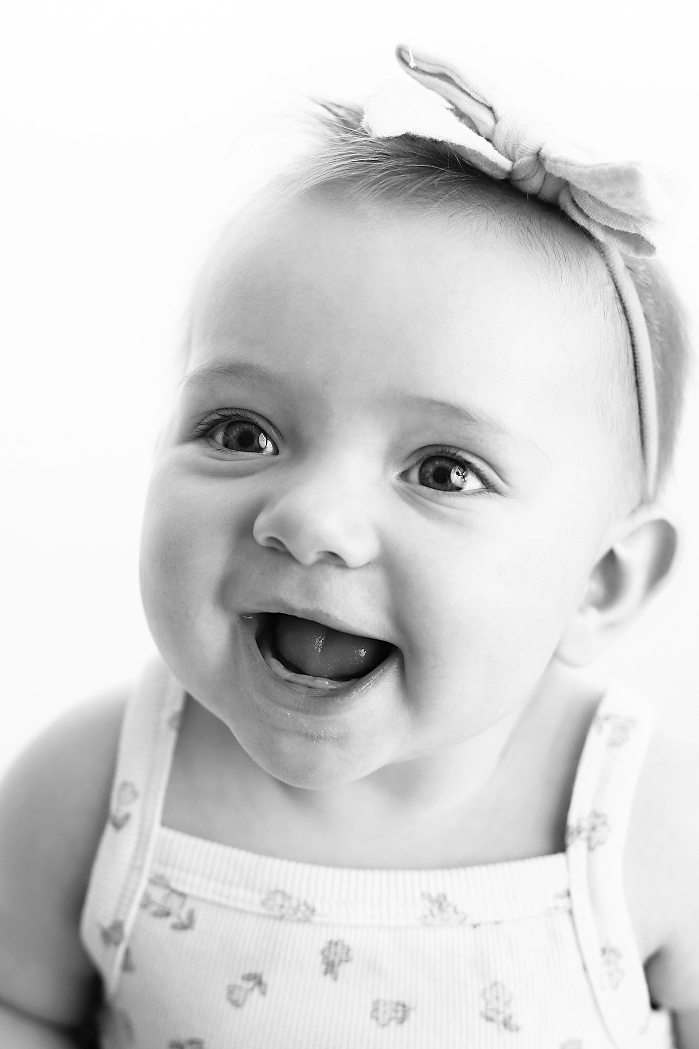 Black and white close-up portrait of a smiling baby girl wearing a bow headband and a floral print tank top. Utah baby photographer