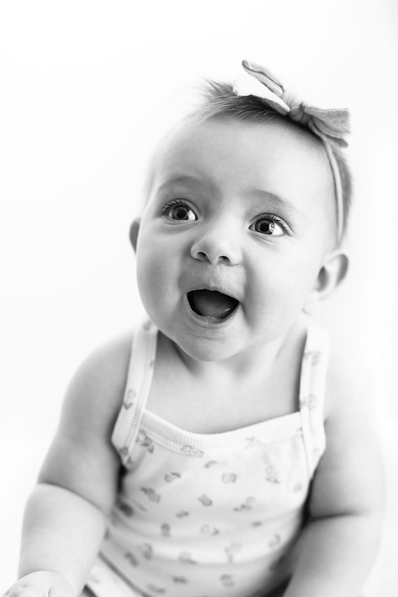 Black and white portrait of a smiling baby with a bow headband wearing a patterned tank top, mouth open in delight.