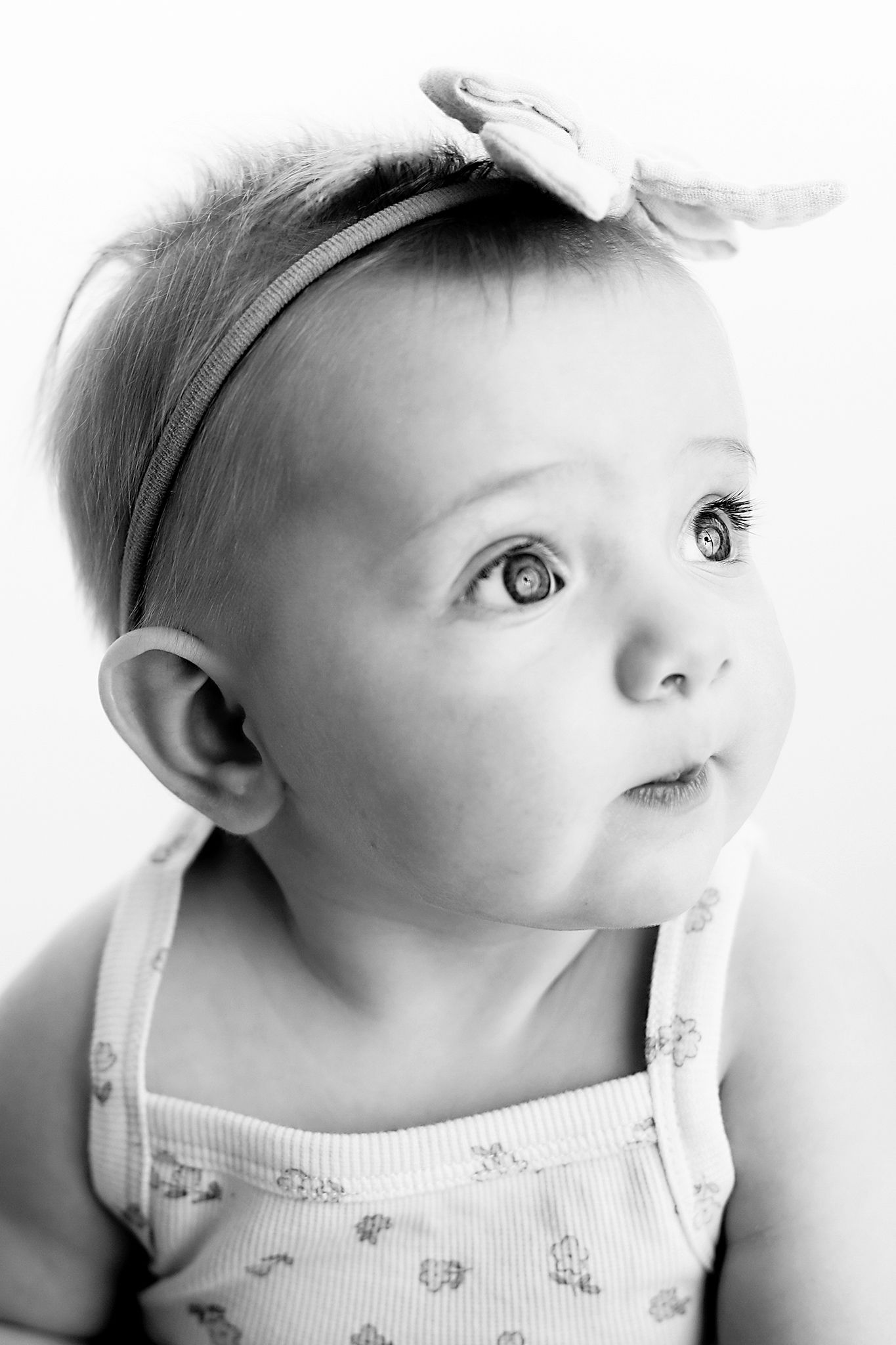 Black and white portrait of a baby girl with a fabric bow headband, wearing a floral tank top and gazing up and to the side.