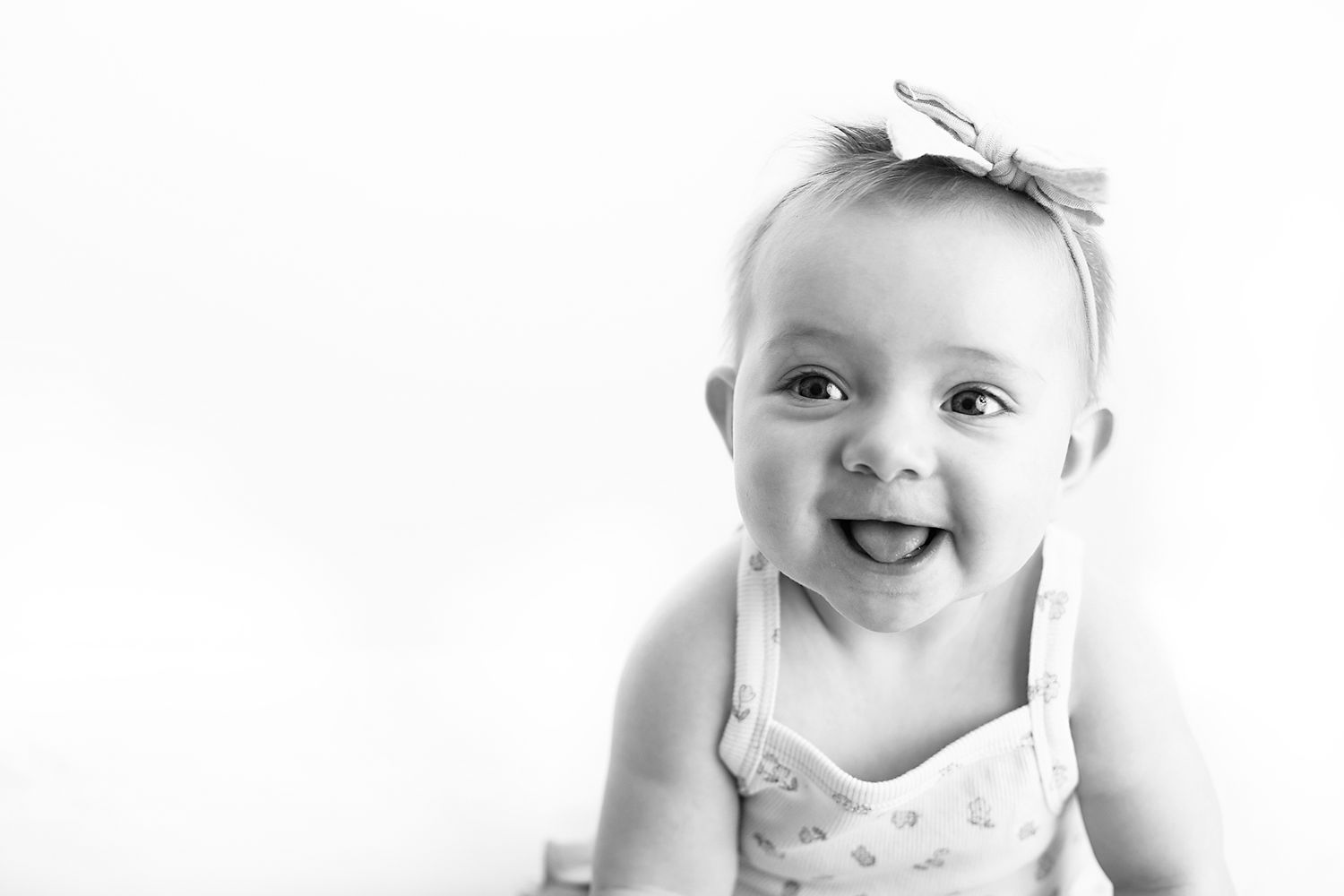 Black and white portrait of a smiling baby girl with a bow headband sticking out her tongue against a clean white background.