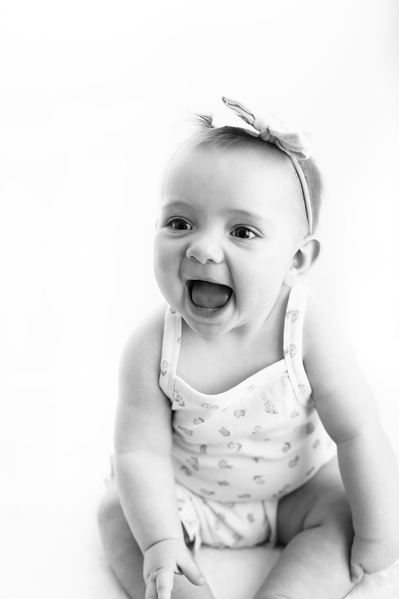 Black and white portrait of a laughing baby girl in a patterned romper and bow headband sitting against a bright white background.