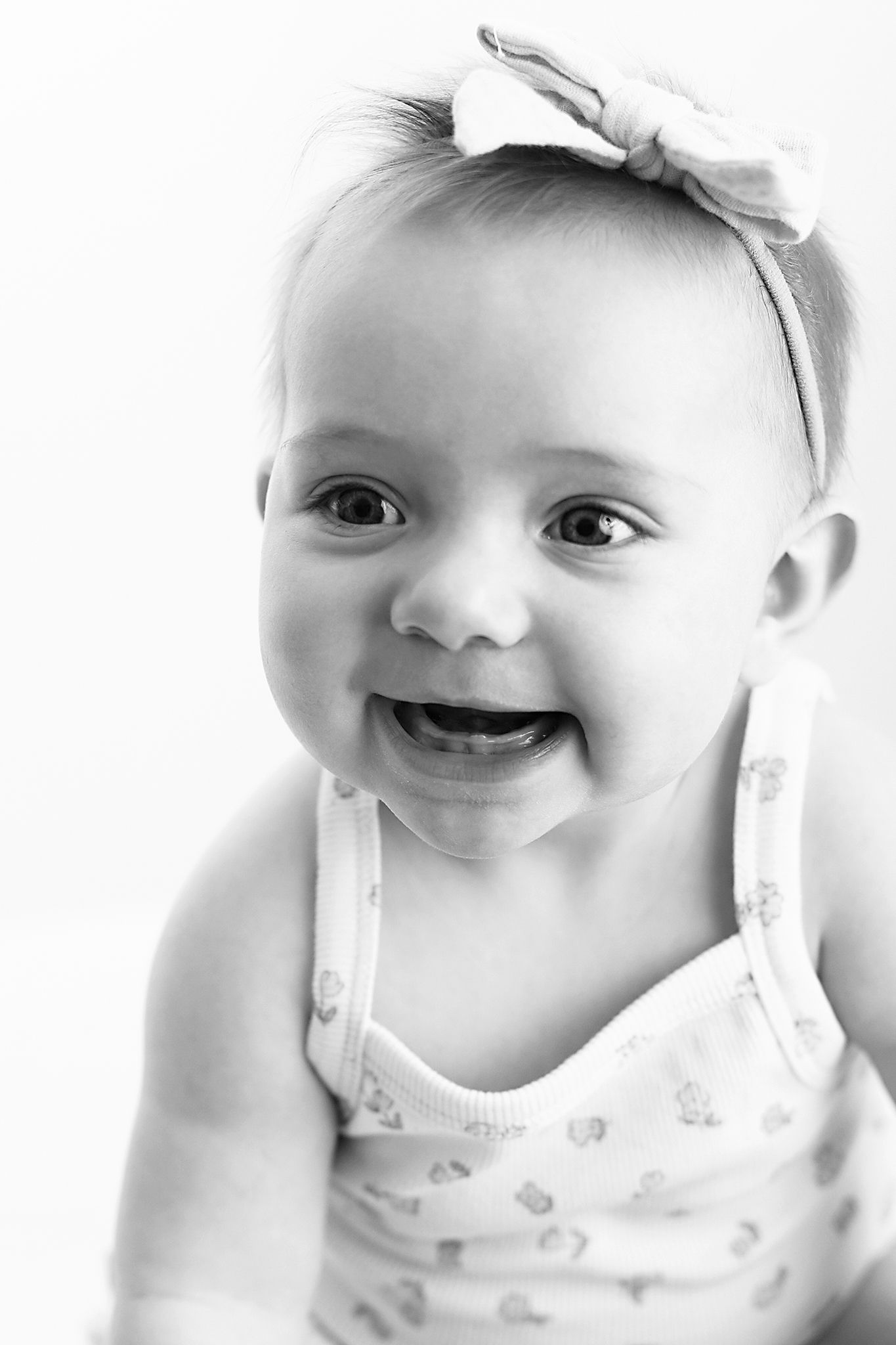 Black and white portrait of a smiling baby girl wearing a bow headband and a delicate patterned tank top.