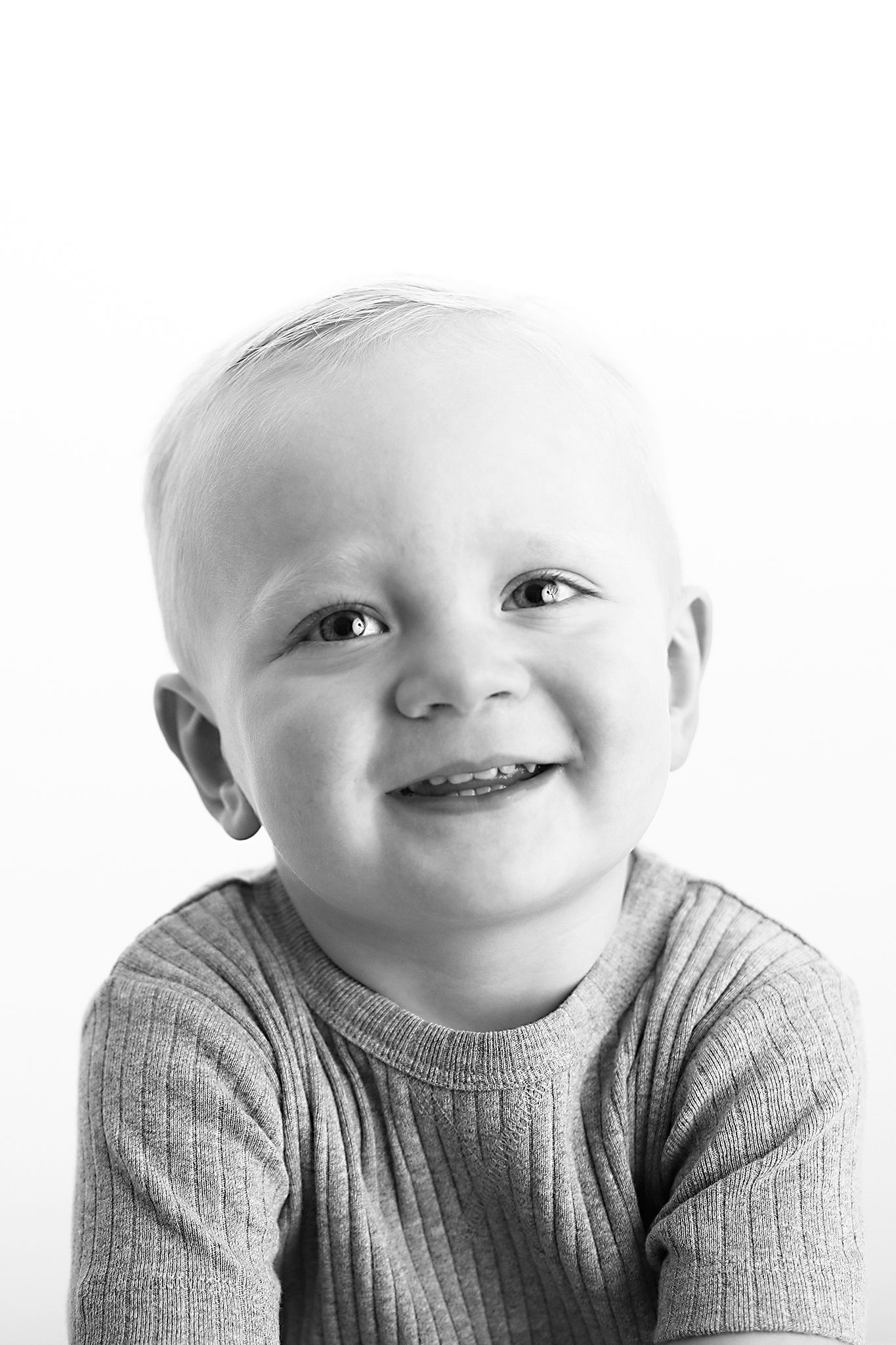 Black and white portrait of a smiling toddler with short light hair wearing a ribbed short-sleeve top against a white background.