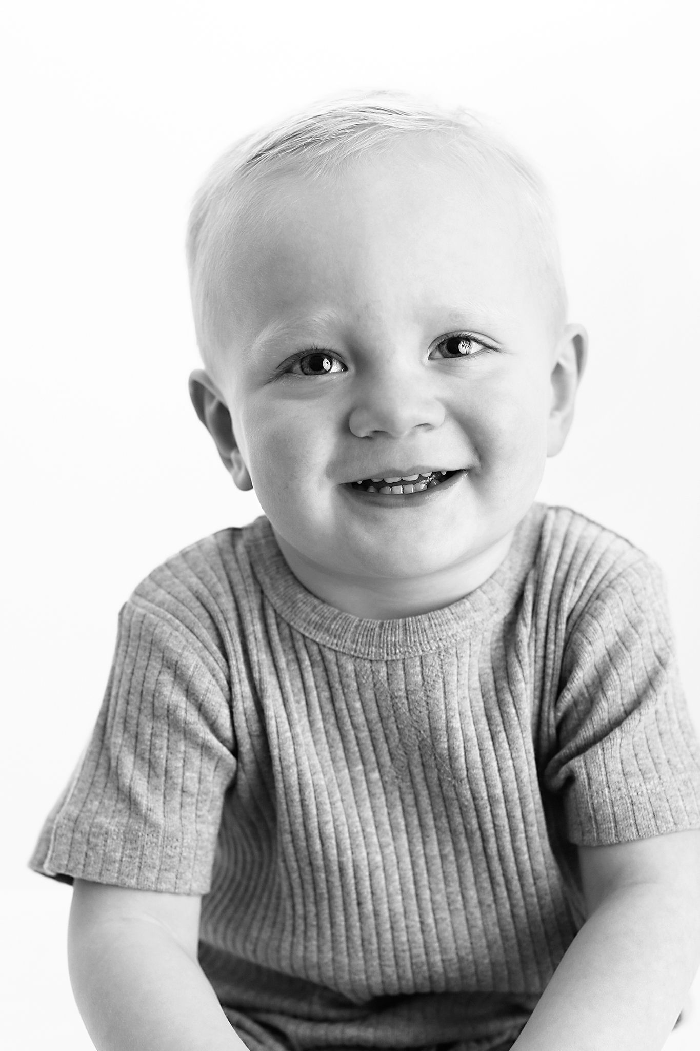 Black and white portrait of a smiling toddler with short hair wearing a ribbed short-sleeve shirt against a plain white background.
