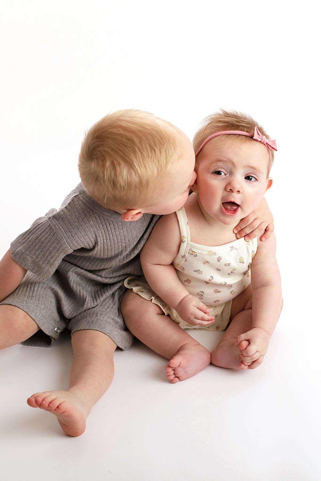Toddler boy in a gray romper kisses the cheek of a baby girl in a pink bow headband against a white background. Utah baby photographer