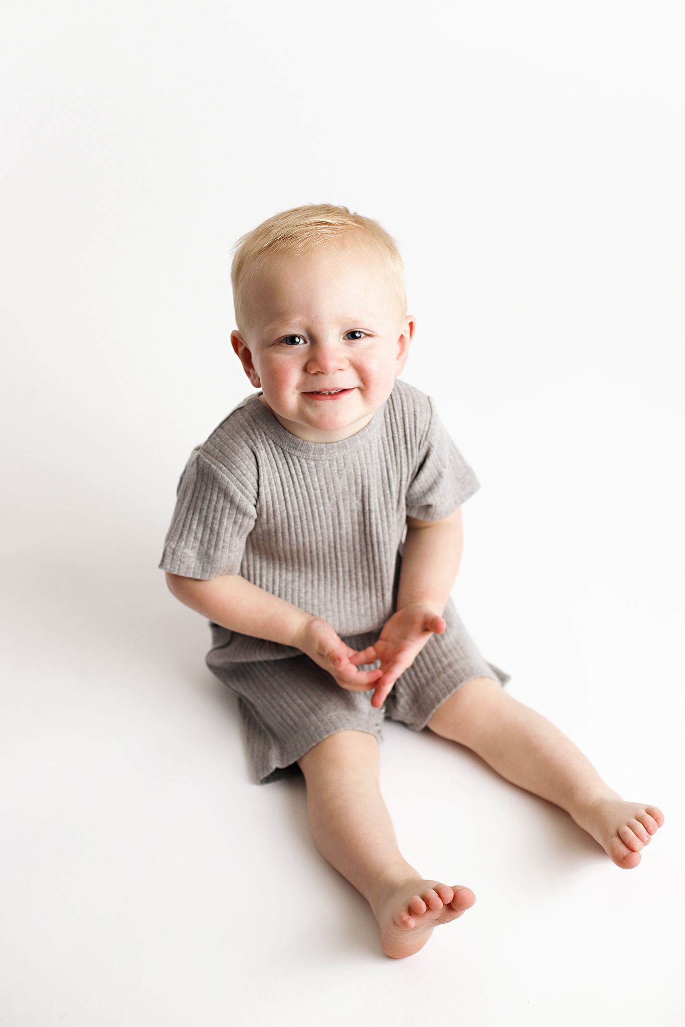 Smiling blond toddler in a gray ribbed short-sleeve romper sits barefoot on a white background with hands together.