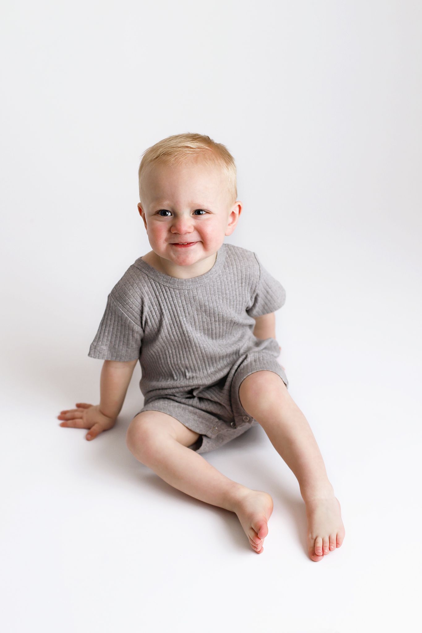 Toddler with blond hair sitting barefoot on a white background, wearing gray ribbed short-sleeve romper and smiling. Utah baby photographer