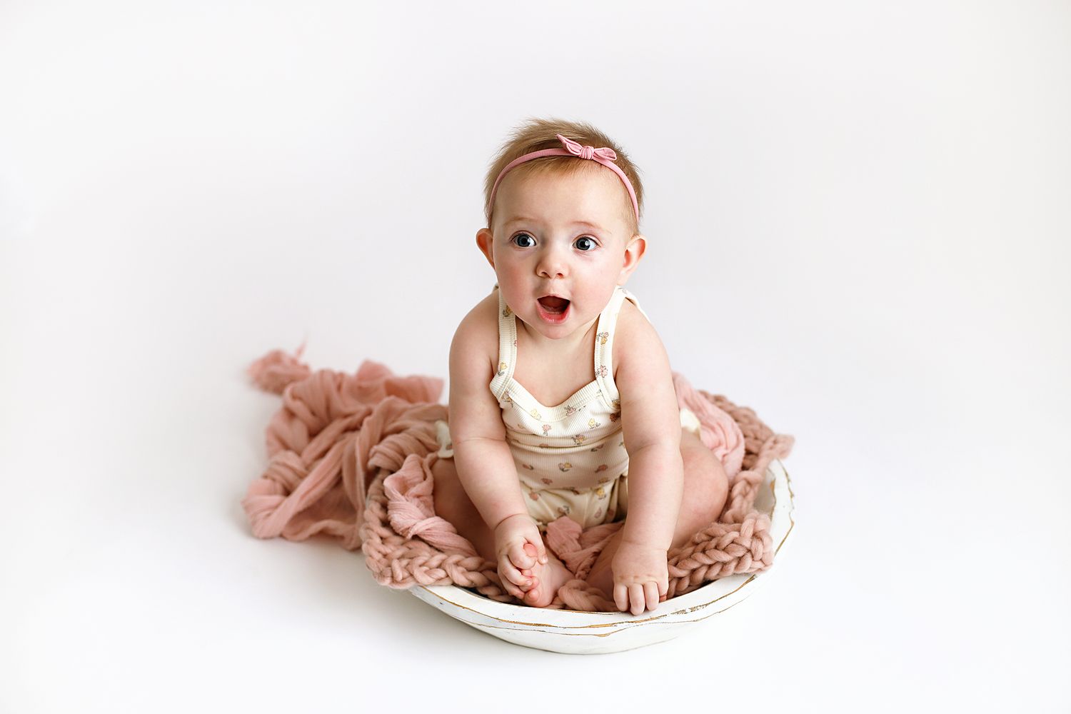 Baby girl in a pink headband and cream outfit sits in a white bowl on a blush knit blanket, gazing sideways with an open mouth.