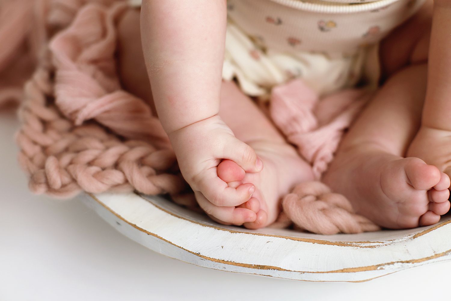 Close-up of a newborn's tiny chubby feet and hands resting in a white wooden bowl with soft pink knit blankets.
