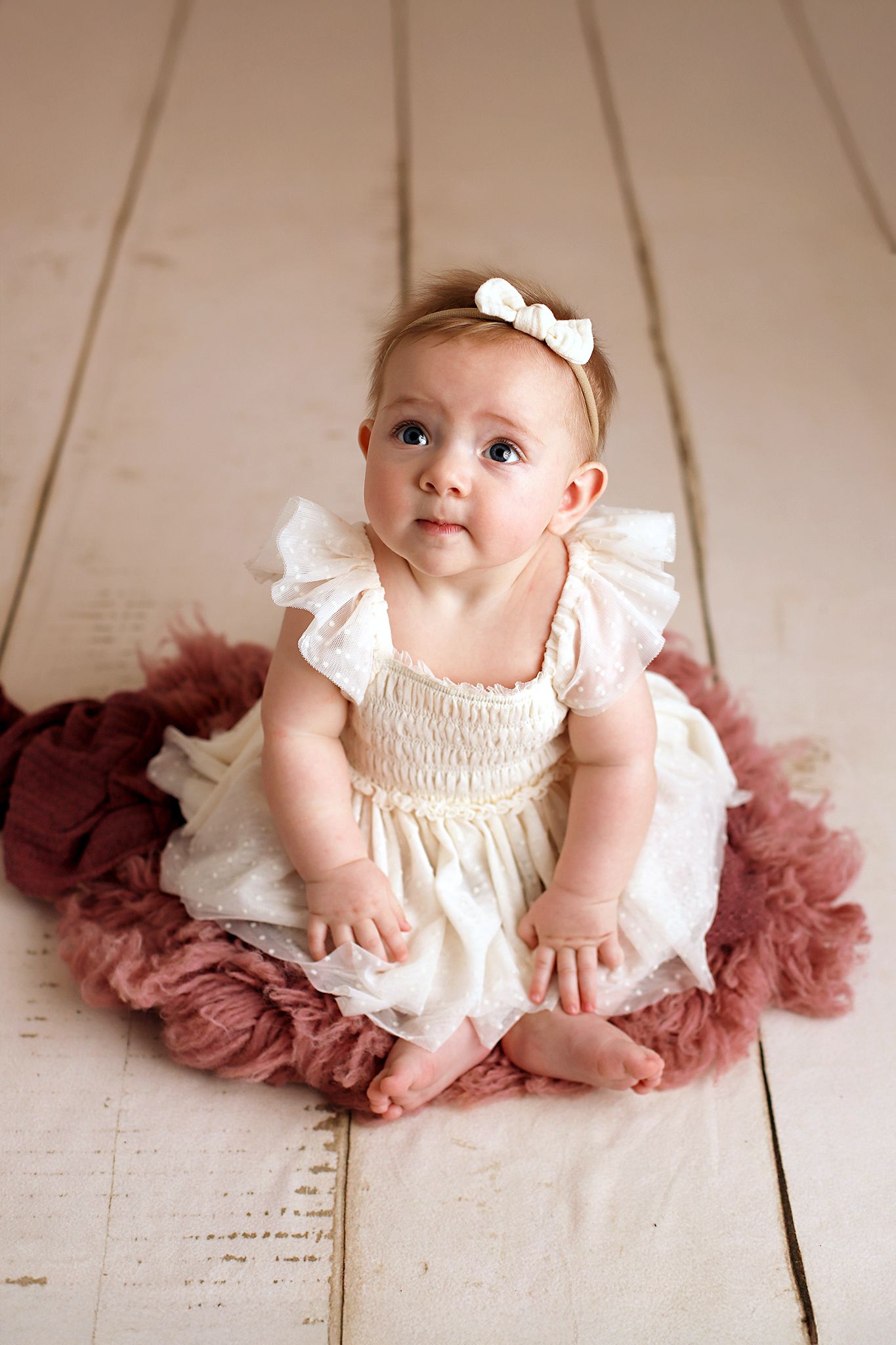 Baby girl in a cream ruffled dress and white bow headband sits on a mauve tulle blanket on a white wooden floor, looking up.