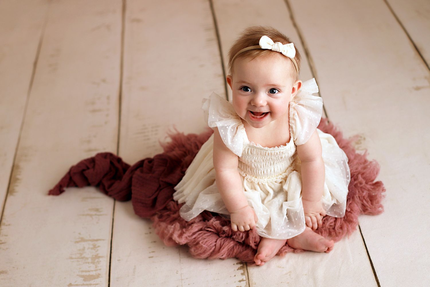Smiling baby girl in a cream tutu dress and white bow headband sitting on a dusty rose ruffled blanket on a white wooden floor.