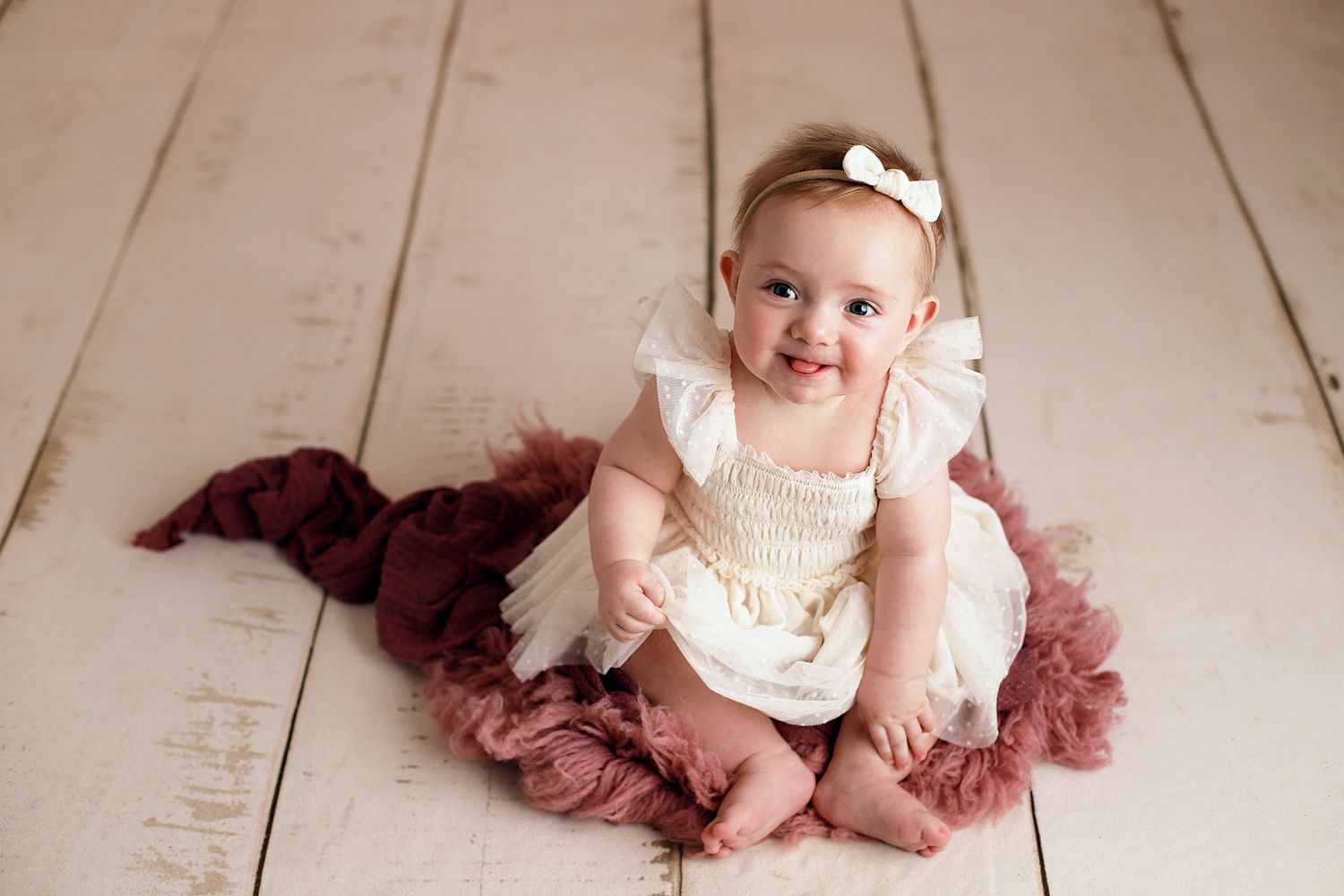 Smiling baby girl in a white lace romper and bow headband sitting on a mauve fluffy blanket over white wooden floor. Utah baby photographer