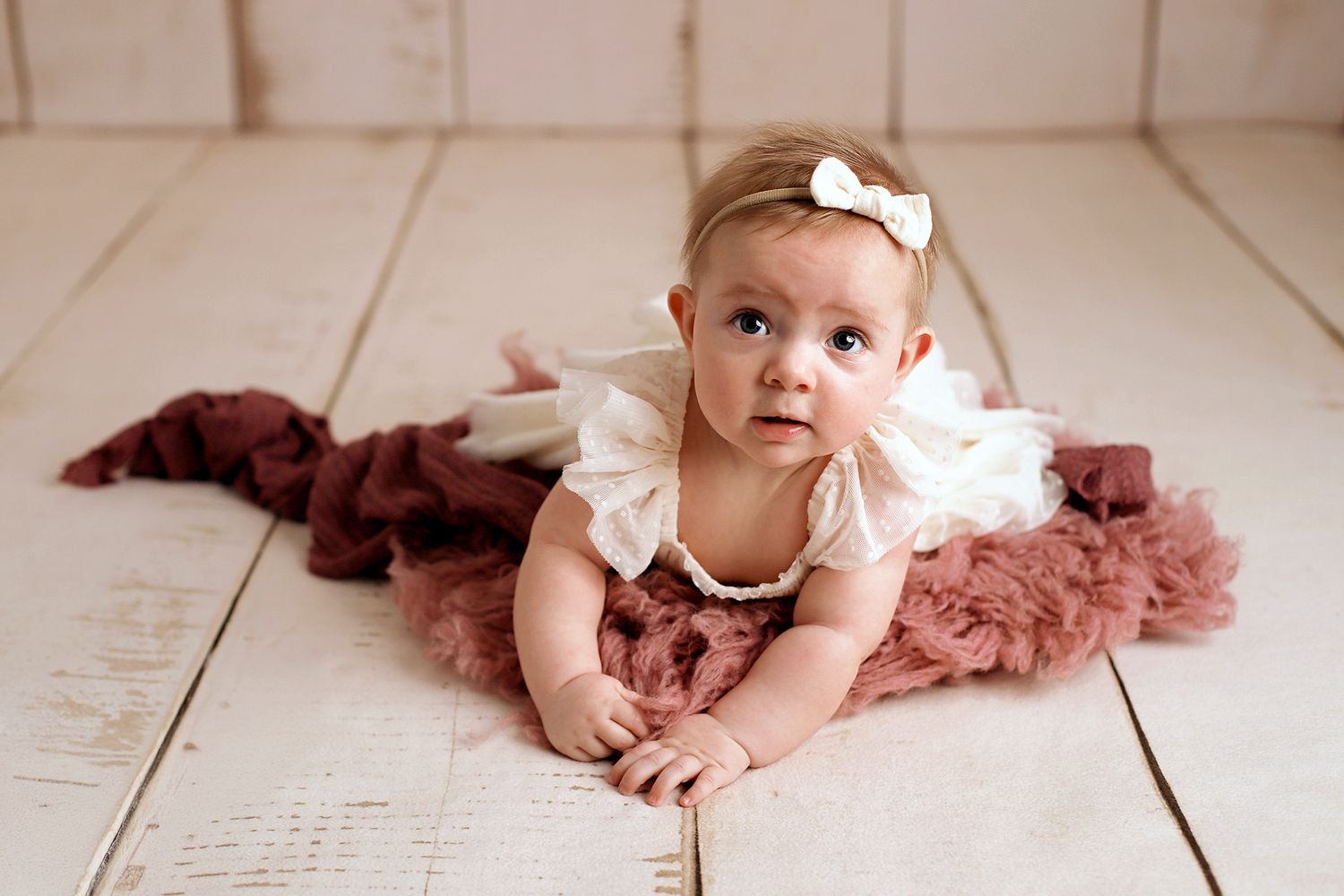 Baby girl in a white bow headband and white lace outfit lying on a burgundy tulle blanket on a whitewashed wooden floor.