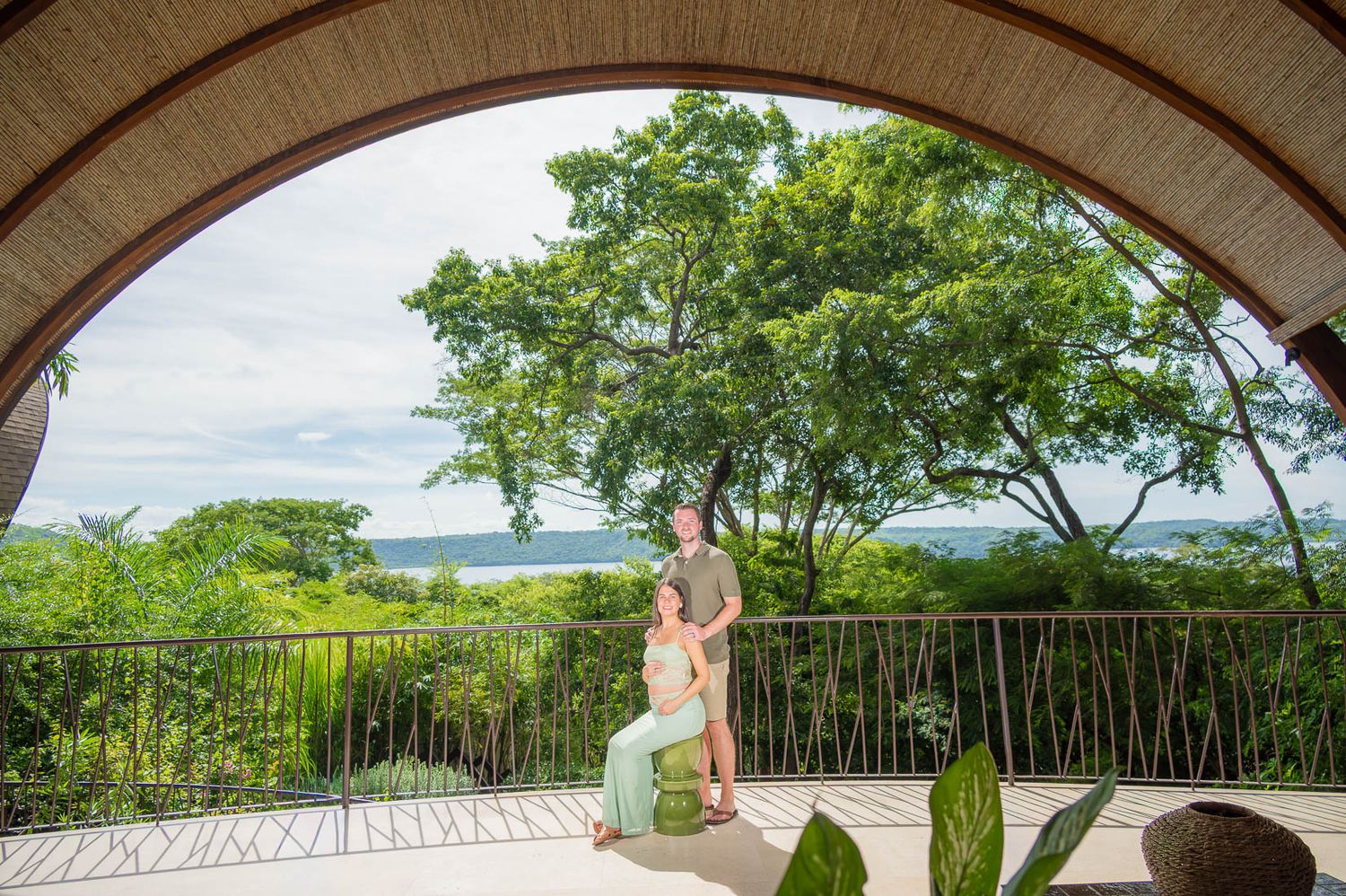 Couple embracing on a covered terrace with a curved archway, overlooking lush tropical trees and a distant ocean view under a cloudy sky.