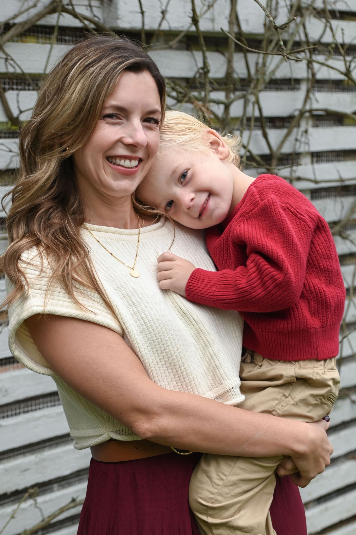Smiling woman in a cream top holding a happy toddler in a red sweater and khaki pants in front of a weathered wooden fence.