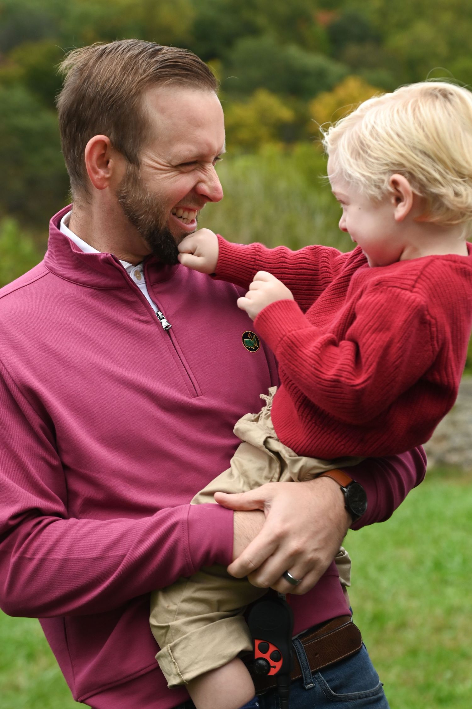 Smiling bearded man in a maroon quarter-zip pullover holds a laughing toddler in a red sweater outdoors with autumn trees in the background.