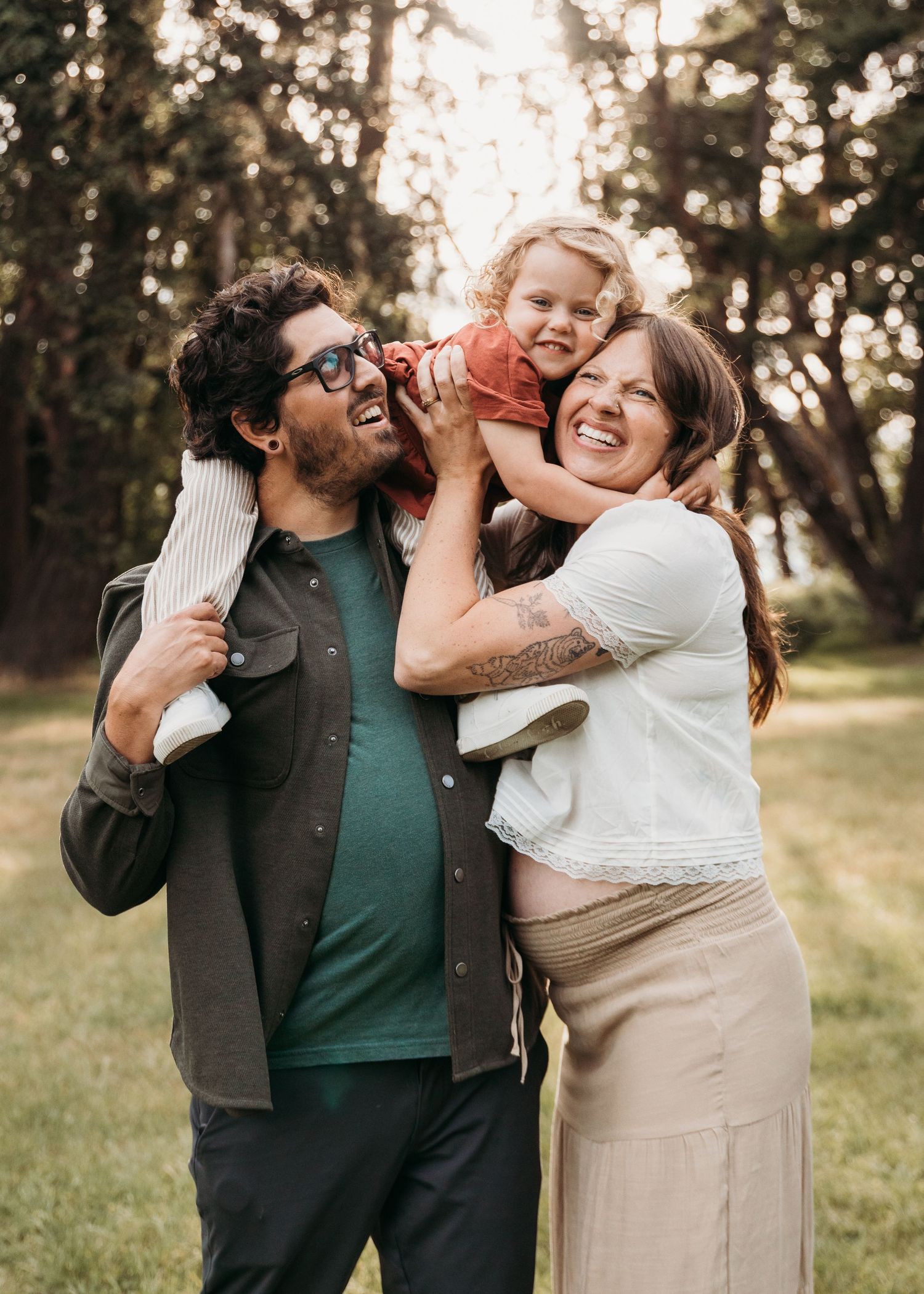 Smiling bearded man in glasses and pregnant woman laugh together outdoors as a toddler sits on his shoulders in a sunny park.