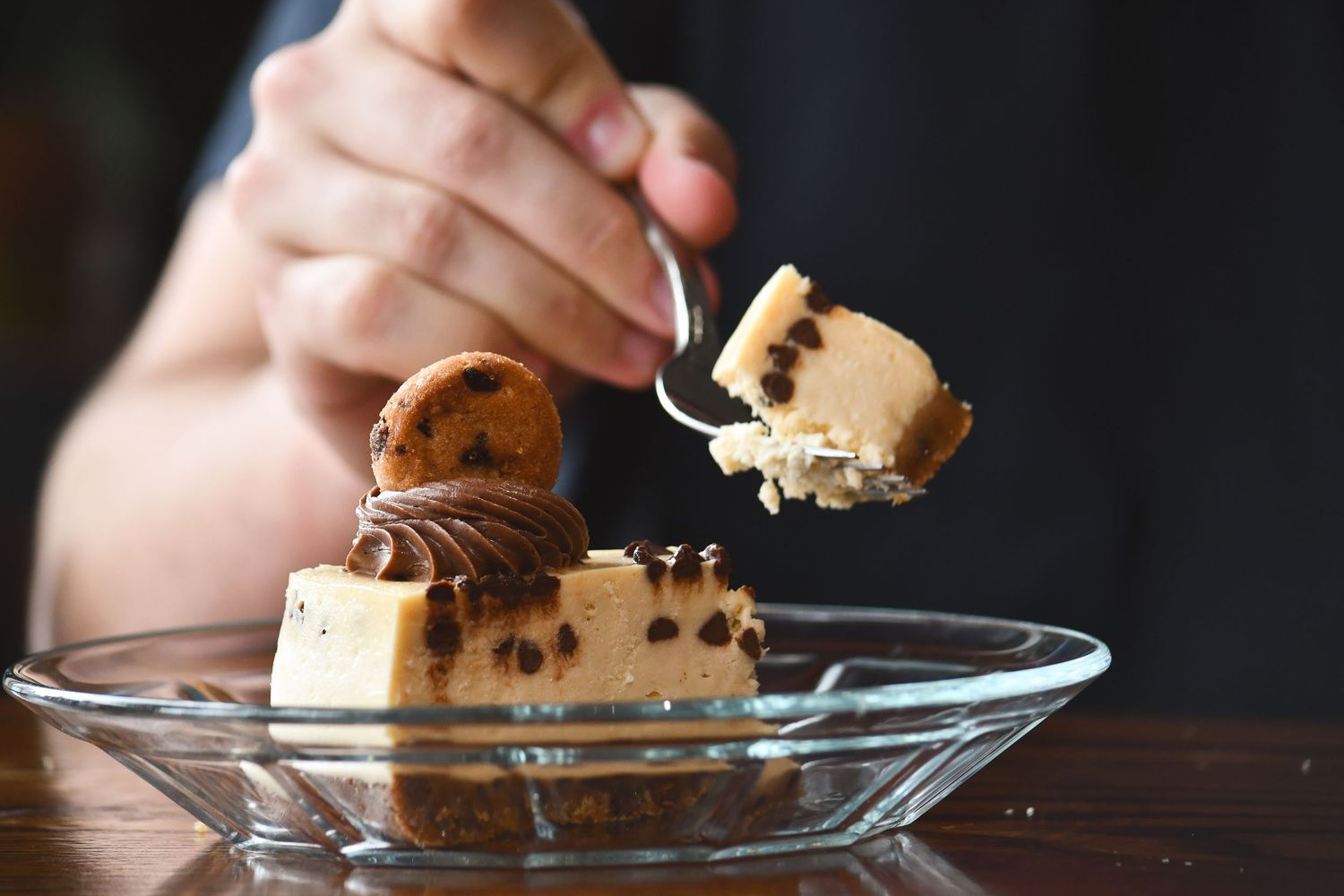 Person eating a slice of chocolate chip cookie dough cheesecake on a glass plate with a spoon.