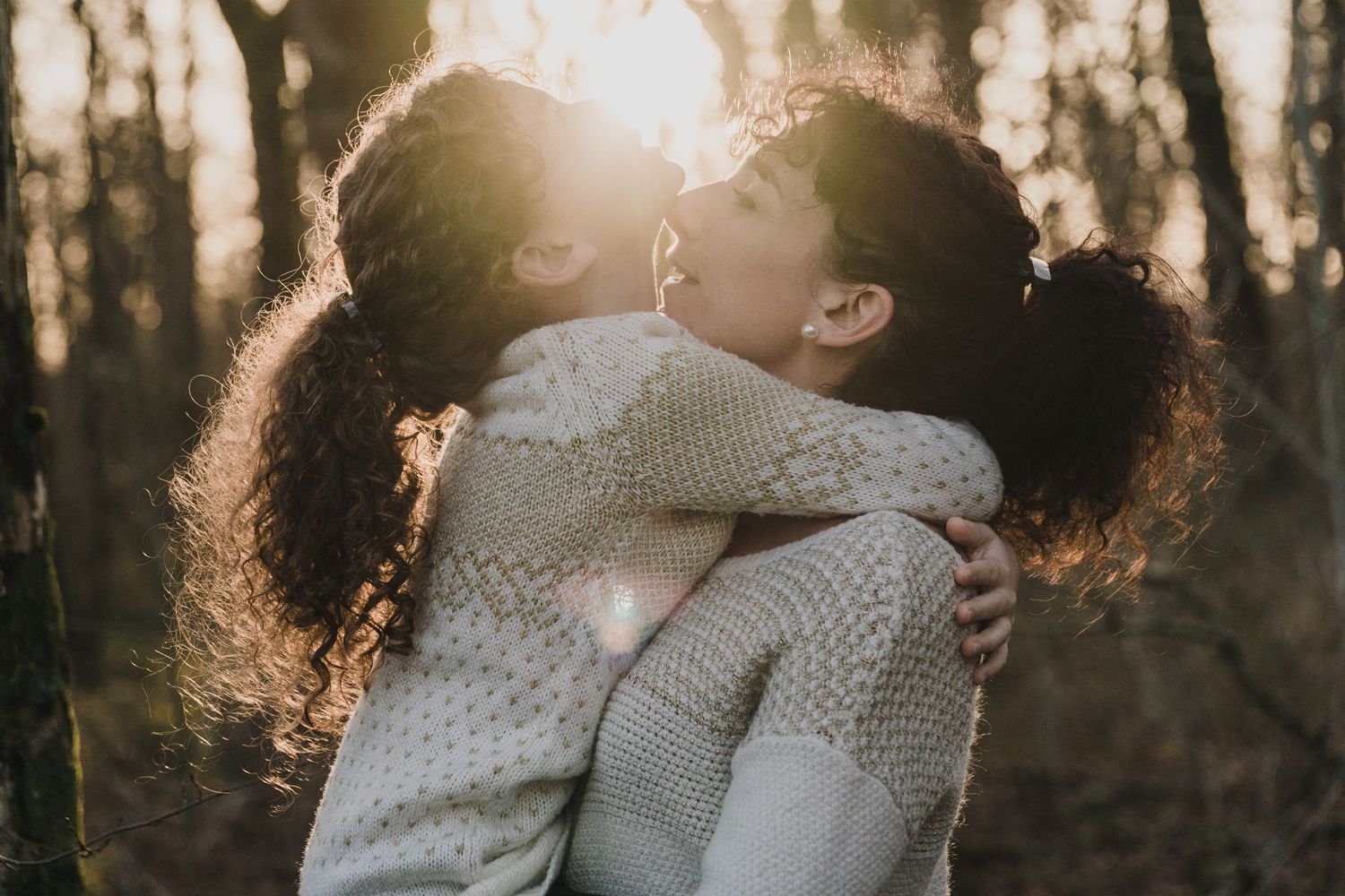 Two girls with curly ponytails embrace and kiss in a forest, warm sunlight filtering through the trees behind them.