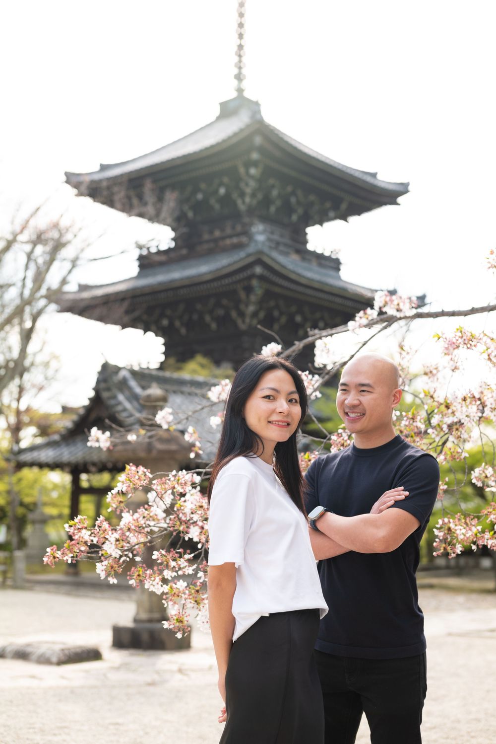 Smiling couple poses beside blooming cherry blossom branches in front of a traditional multi-tiered Japanese pagoda at a temple.