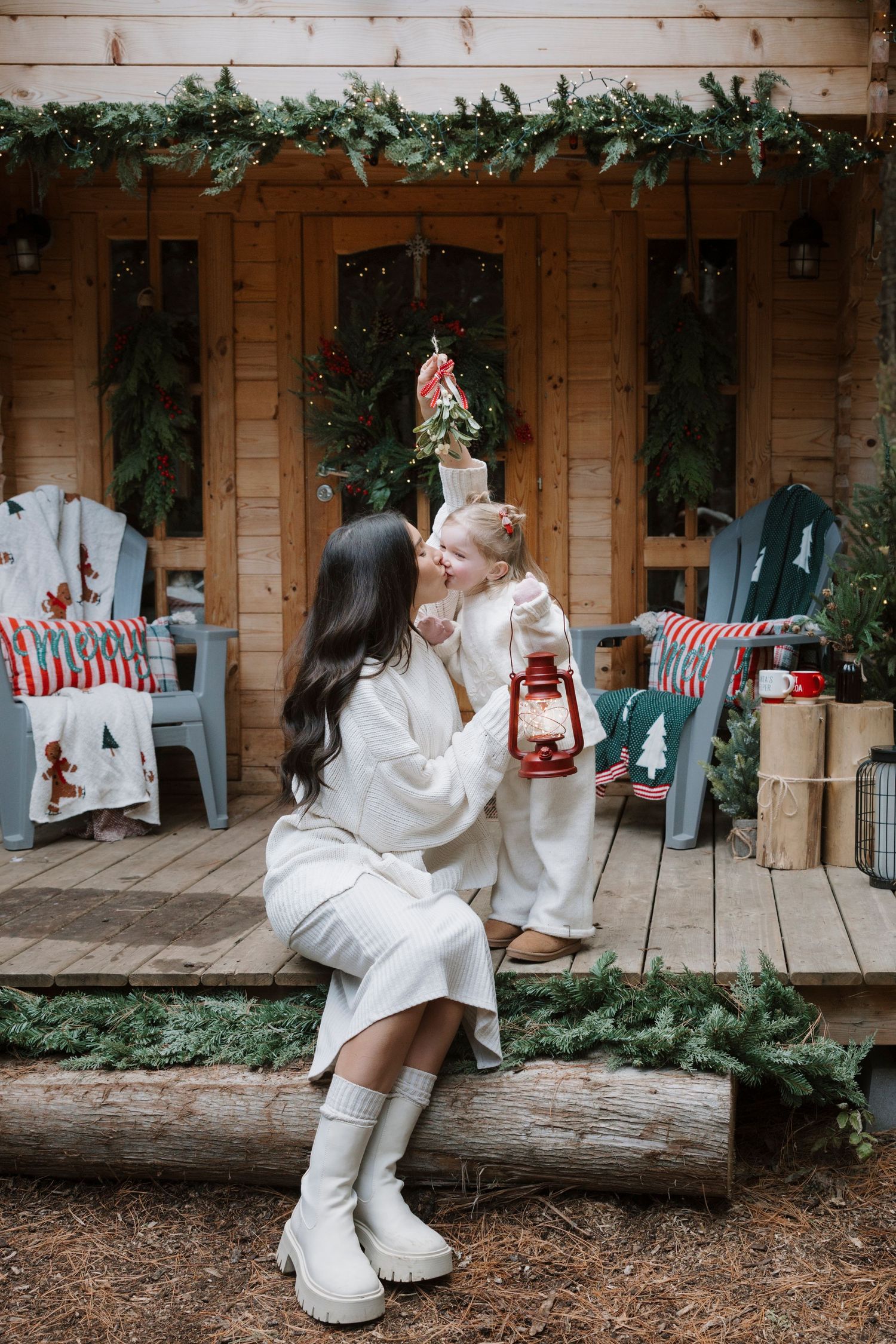 Mother and daughter in white outfits share a kiss holding red lantern on festive Christmas cabin porch.
