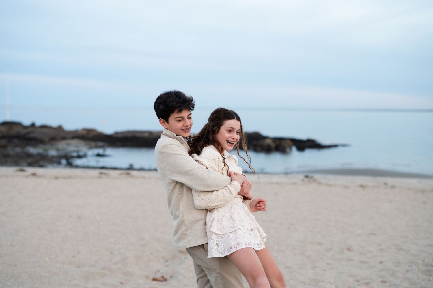 Siblings laughing and dancing together on a sandy beach at dusk, dressed in coordinating cream outfits.