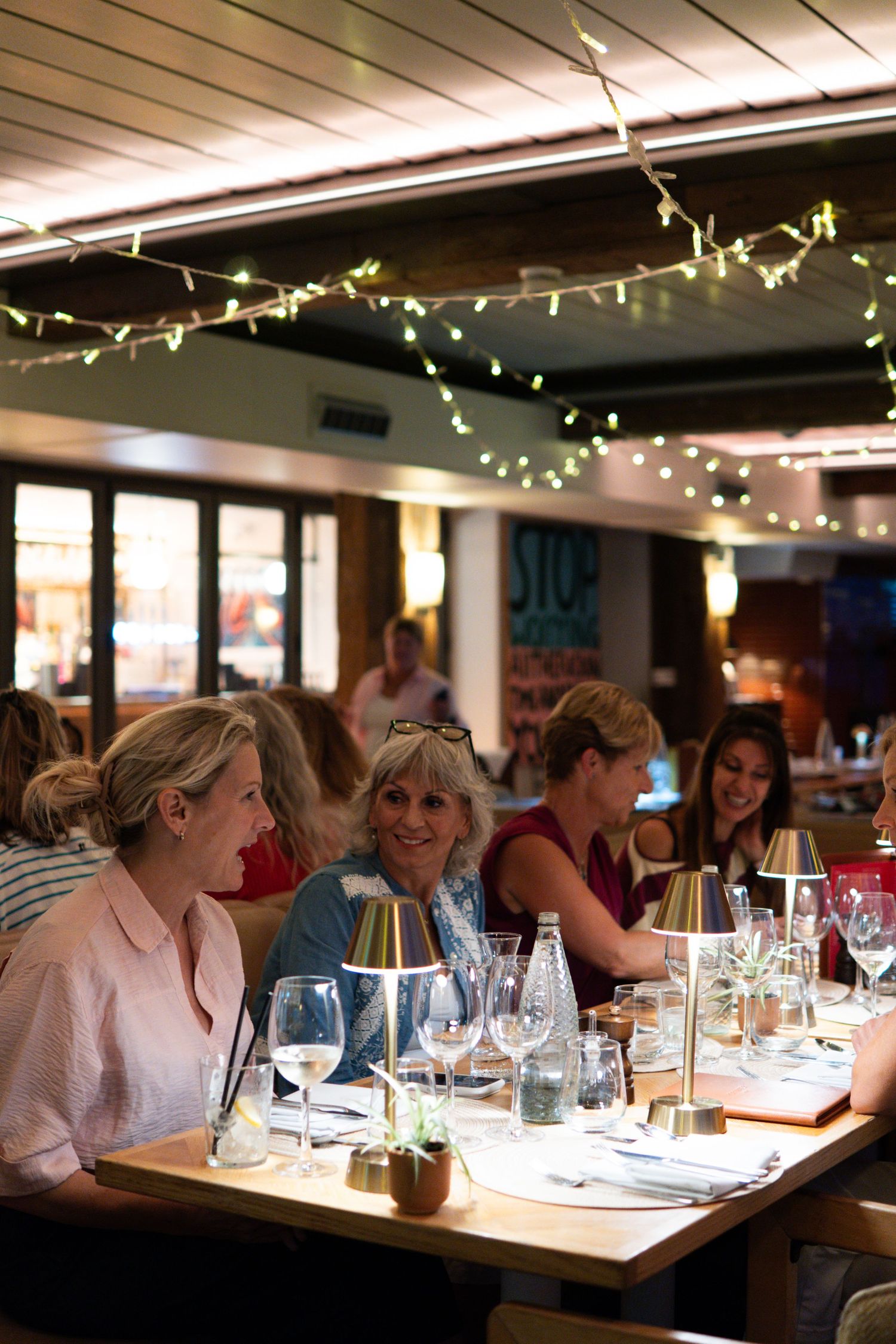 Guests dining at a warmly lit restaurant with fairy lights, long tables, and an inviting evening atmosphere.