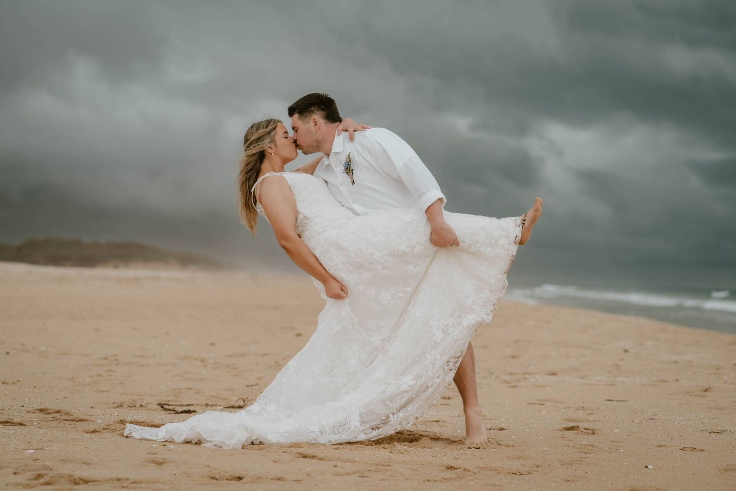 Couple sharing a romantic dip kiss on a sandy beach in wedding attire with dramatic stormy skies behind them.