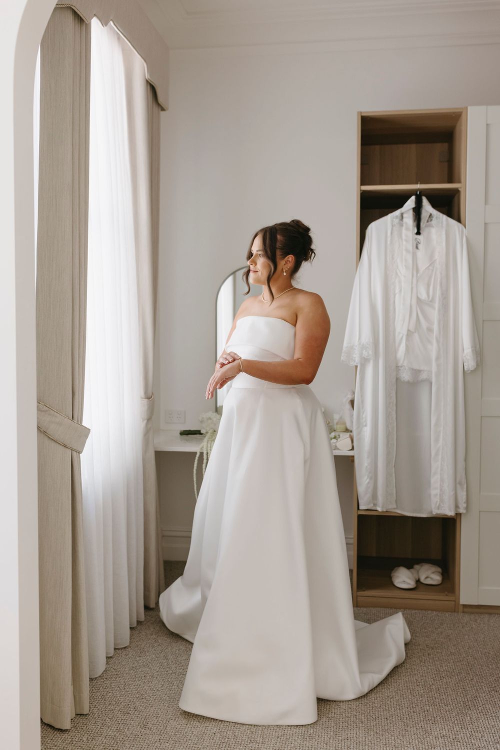 Bride in strapless white wedding gown standing by window with robe hanging in wardrobe behind her.