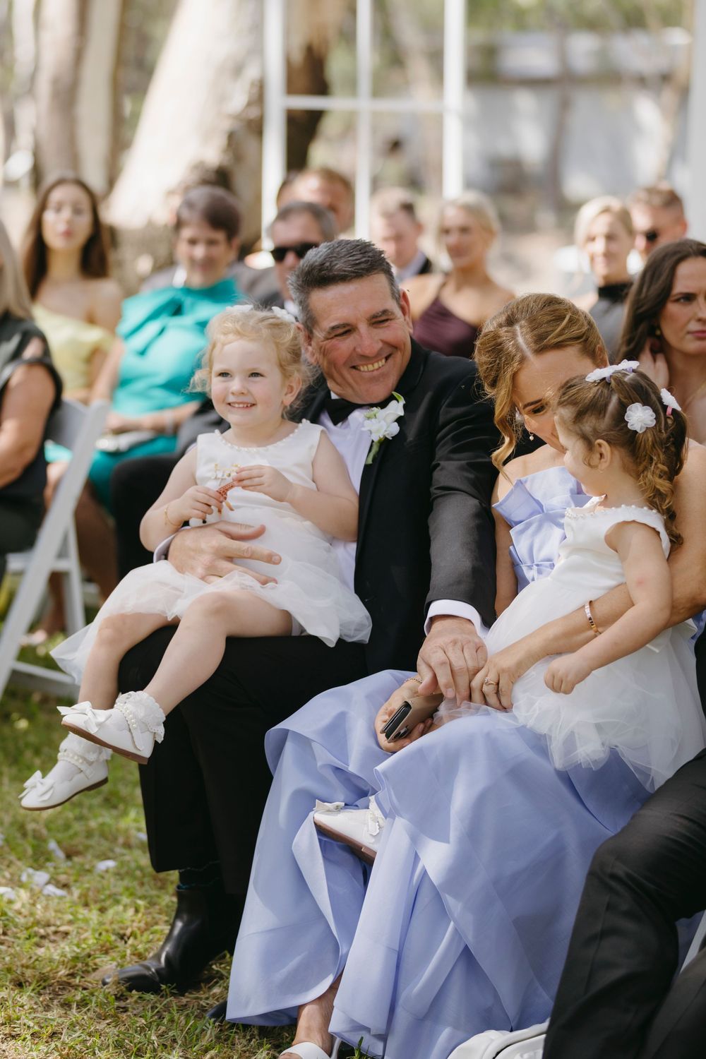Smiling man in tuxedo holds two young flower girls in white dresses at outdoor wedding ceremony.