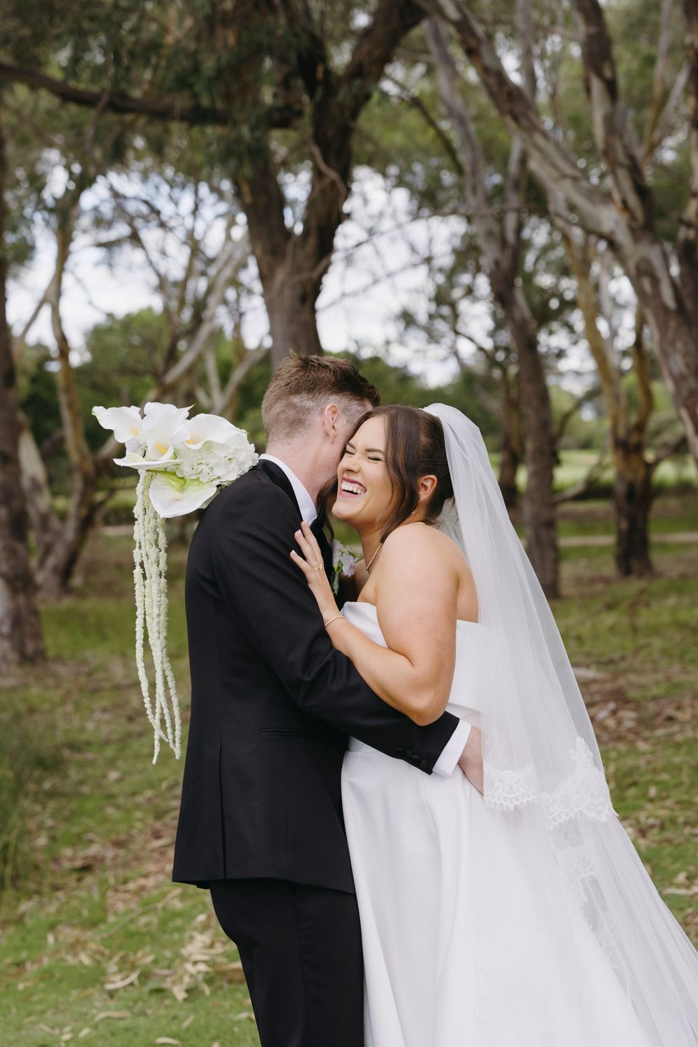 Laughing bride in white gown embraces groom in black suit among trees holding white bouquet.