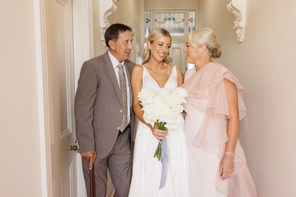 Bride in white gown holding bouquet stands smiling with her parents in an elegant hallway.