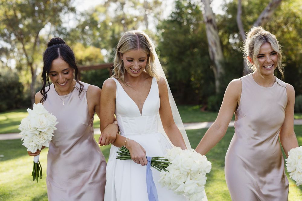 Bride in white gown walks with two bridesmaids in blush dresses carrying white floral bouquets outdoors.