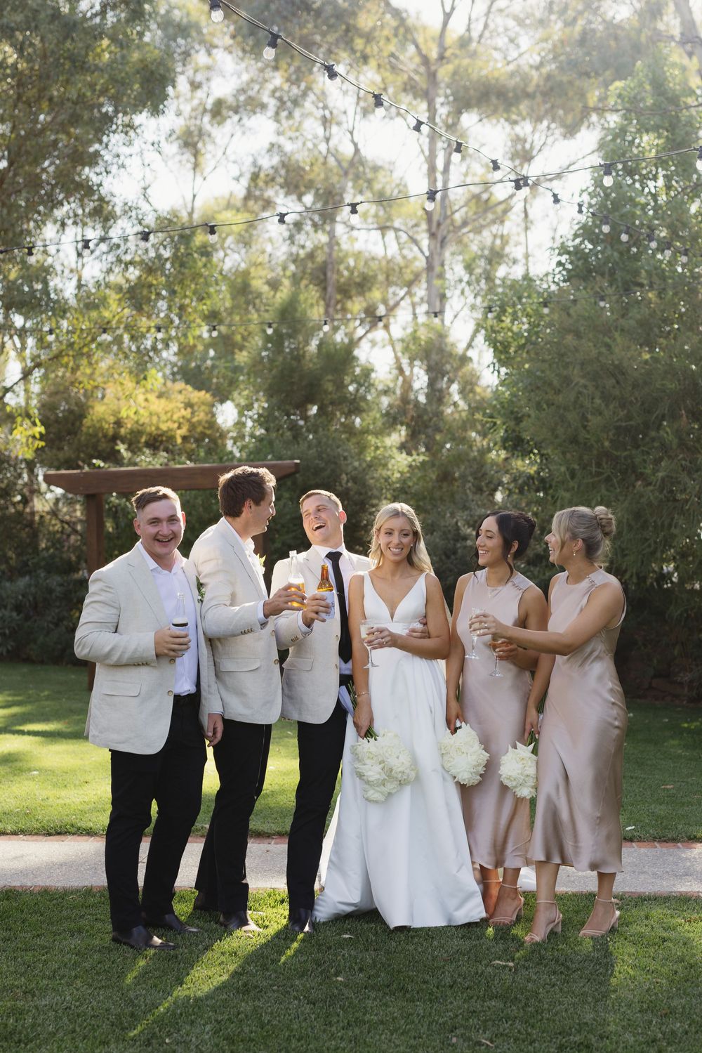 Wedding party laughing and toasting with champagne on a green lawn beneath string lights.