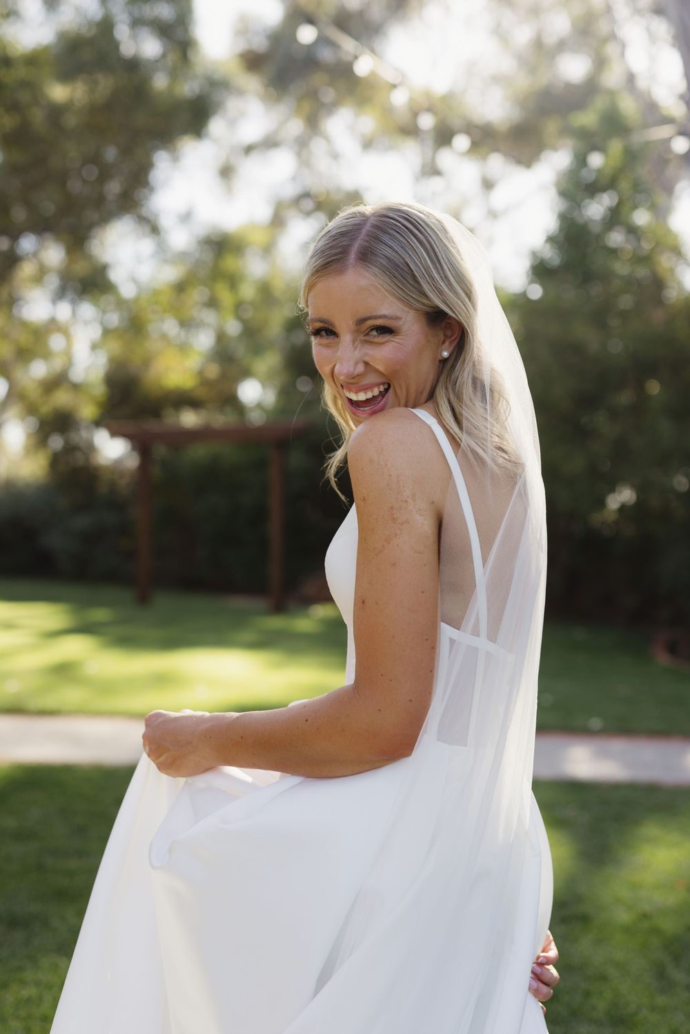 Radiant bride in white gown and veil laughing while holding her dress in a lush garden.