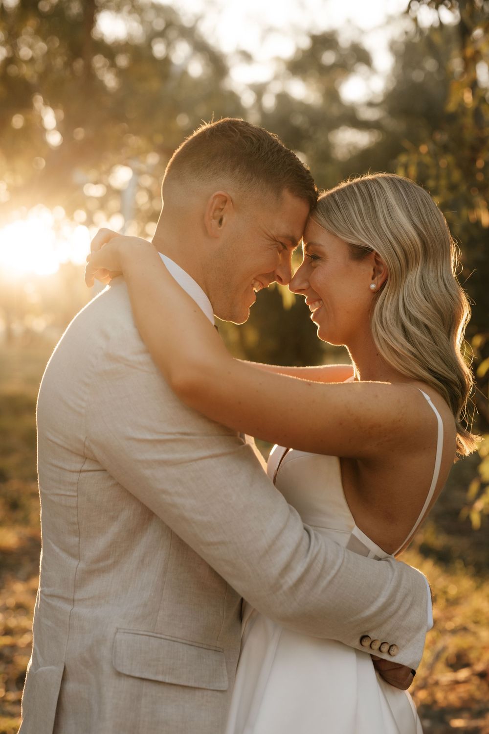 Bride and groom embrace forehead to forehead smiling in golden sunset light among trees.