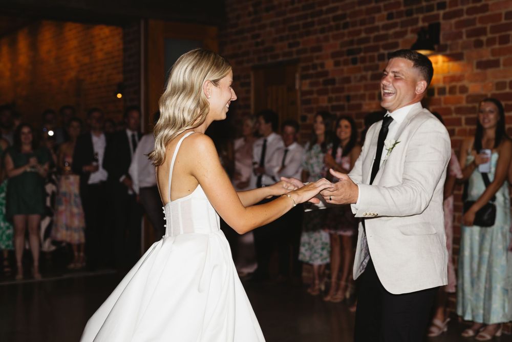 Bride and groom share a joyful first dance holding hands in a warmly lit brick venue.
