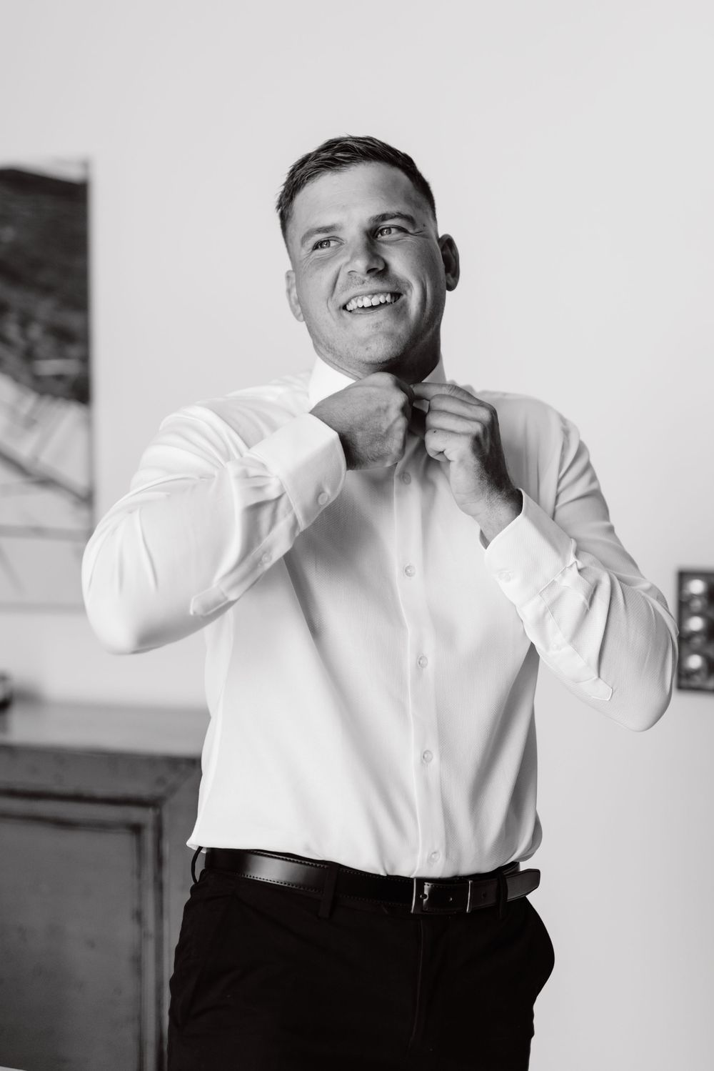 Smiling groom in white dress shirt adjusts collar while getting ready for wedding day.