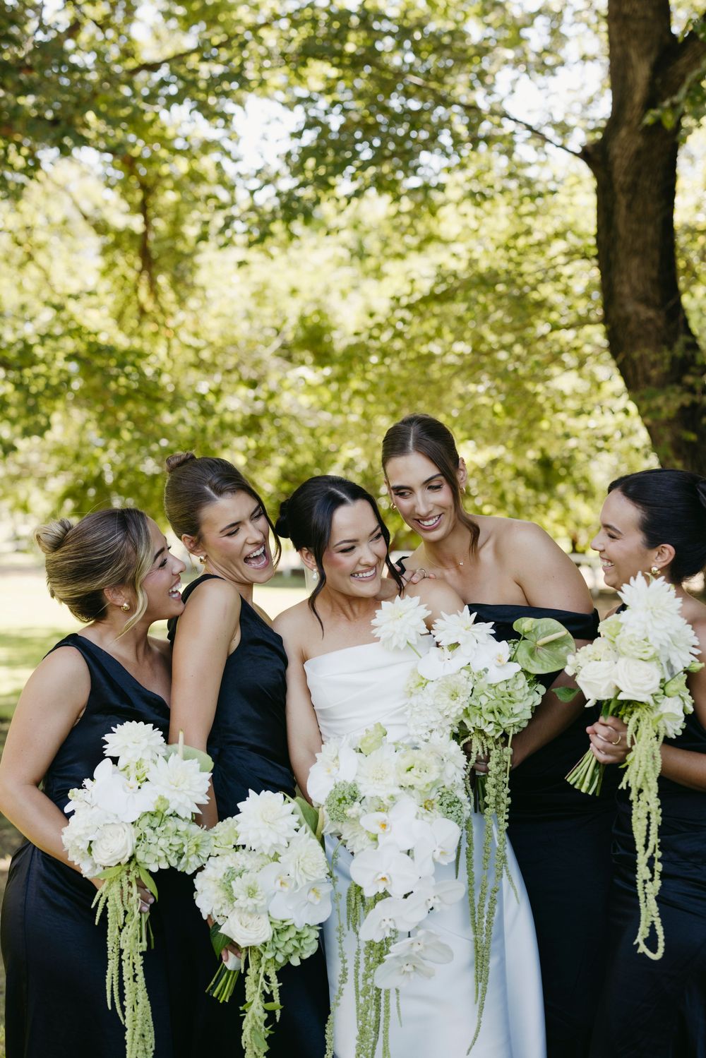 Bride with bridesmaids in black dresses laughing together outdoors holding white floral bouquets.