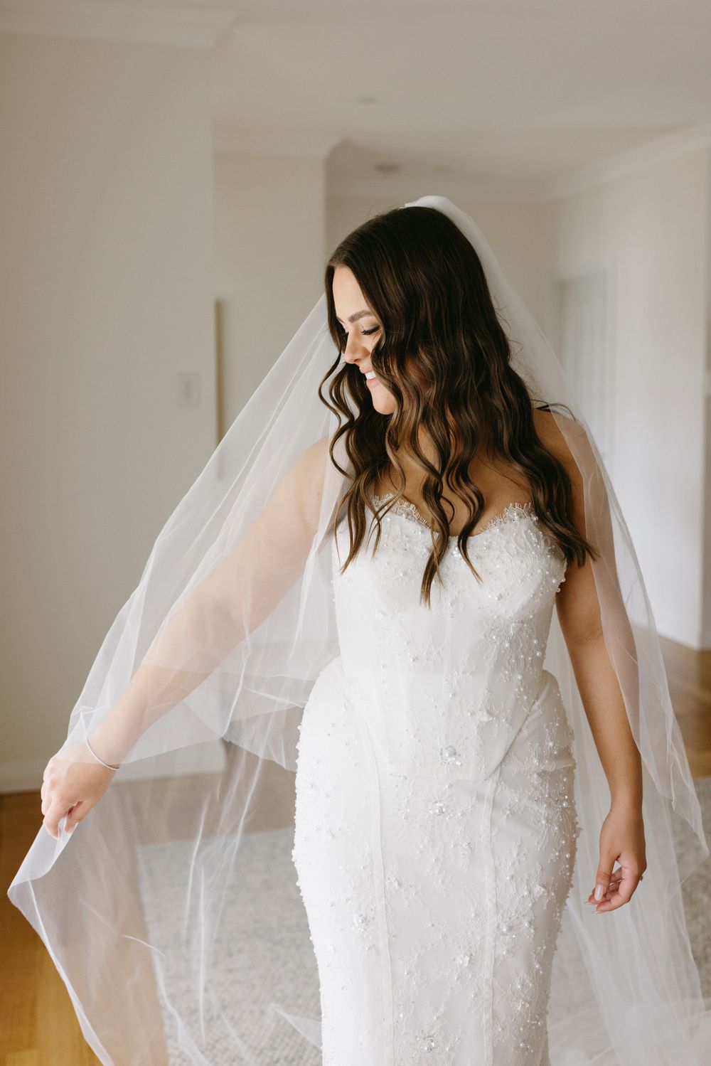 Brunette bride in pearl-embellished white gown holds flowing veil indoors before wedding ceremony.