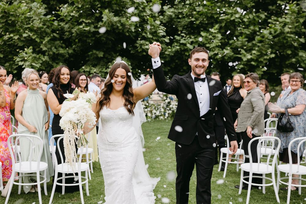 Newlyweds walk down aisle celebrating as guests throw white confetti at outdoor garden wedding.