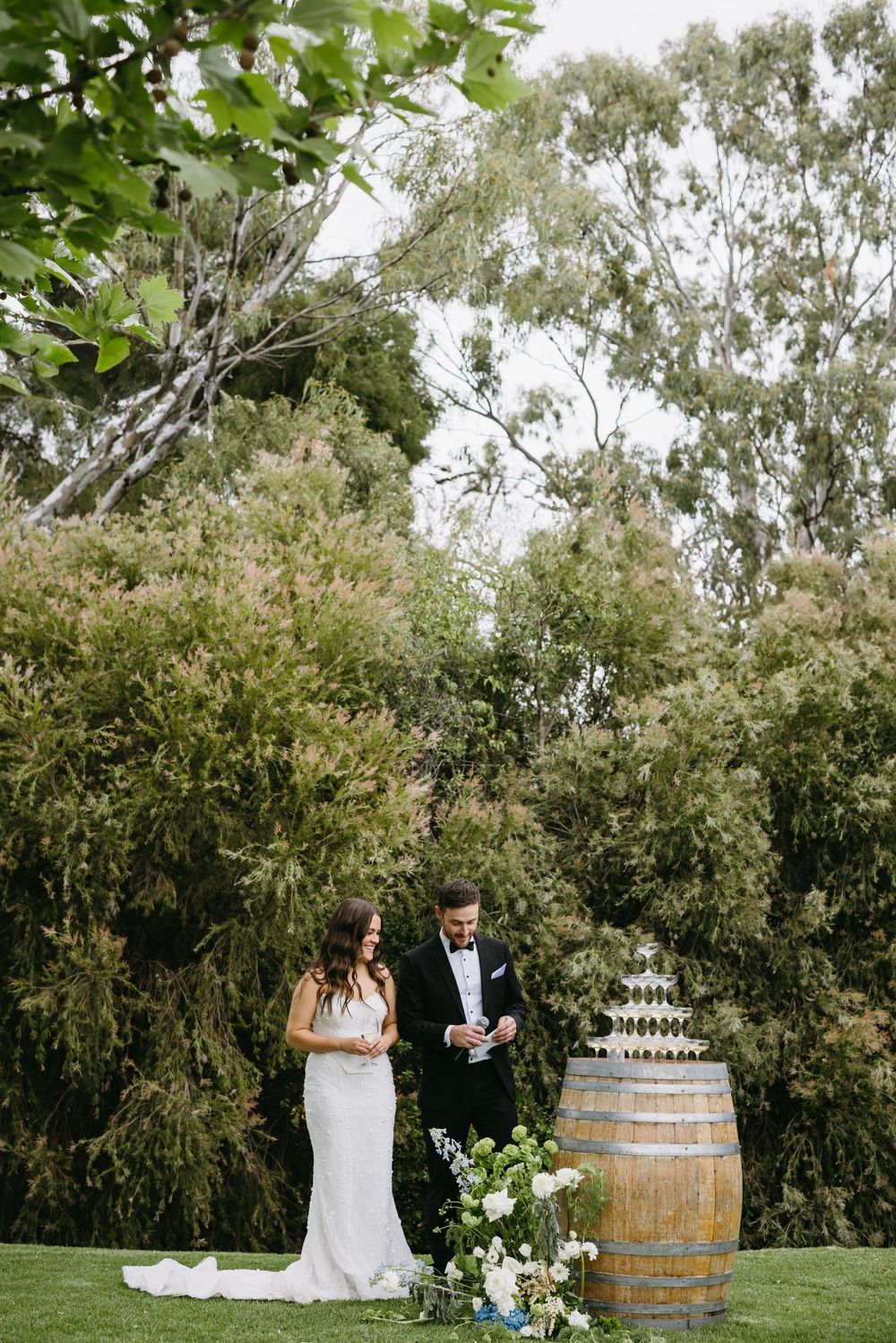 Bride and groom share champagne beside rustic wooden barrel with white floral arrangements outdoors.