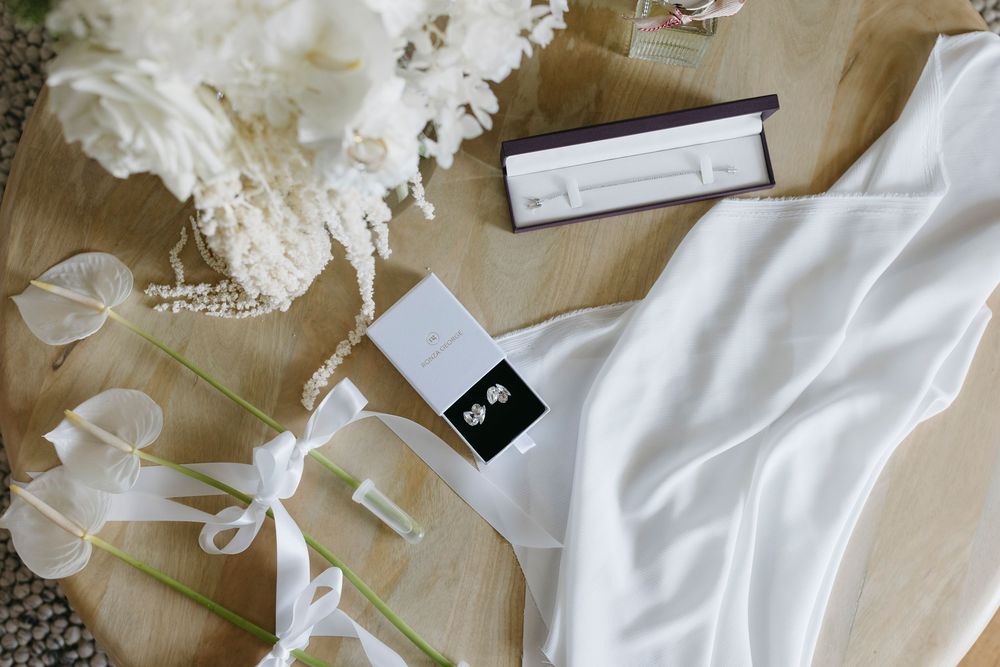Elegant bridal flat lay with white flowers, pearl jewelry, earrings in box, and silk fabric on wooden surface.