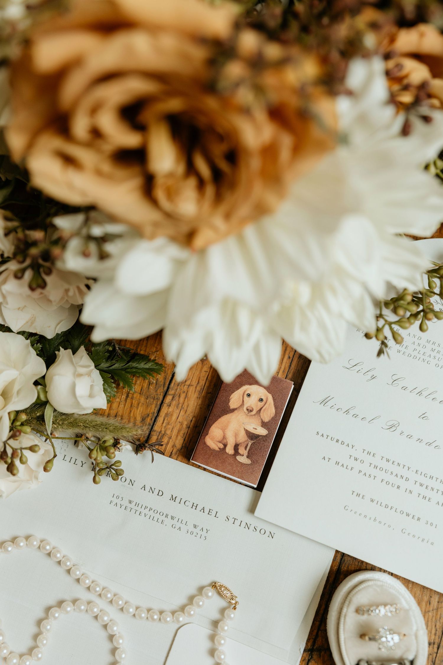 Elegant wedding flat lay with floral bouquet, pearl necklace, invitation suite, and a dachshund illustration on wood.