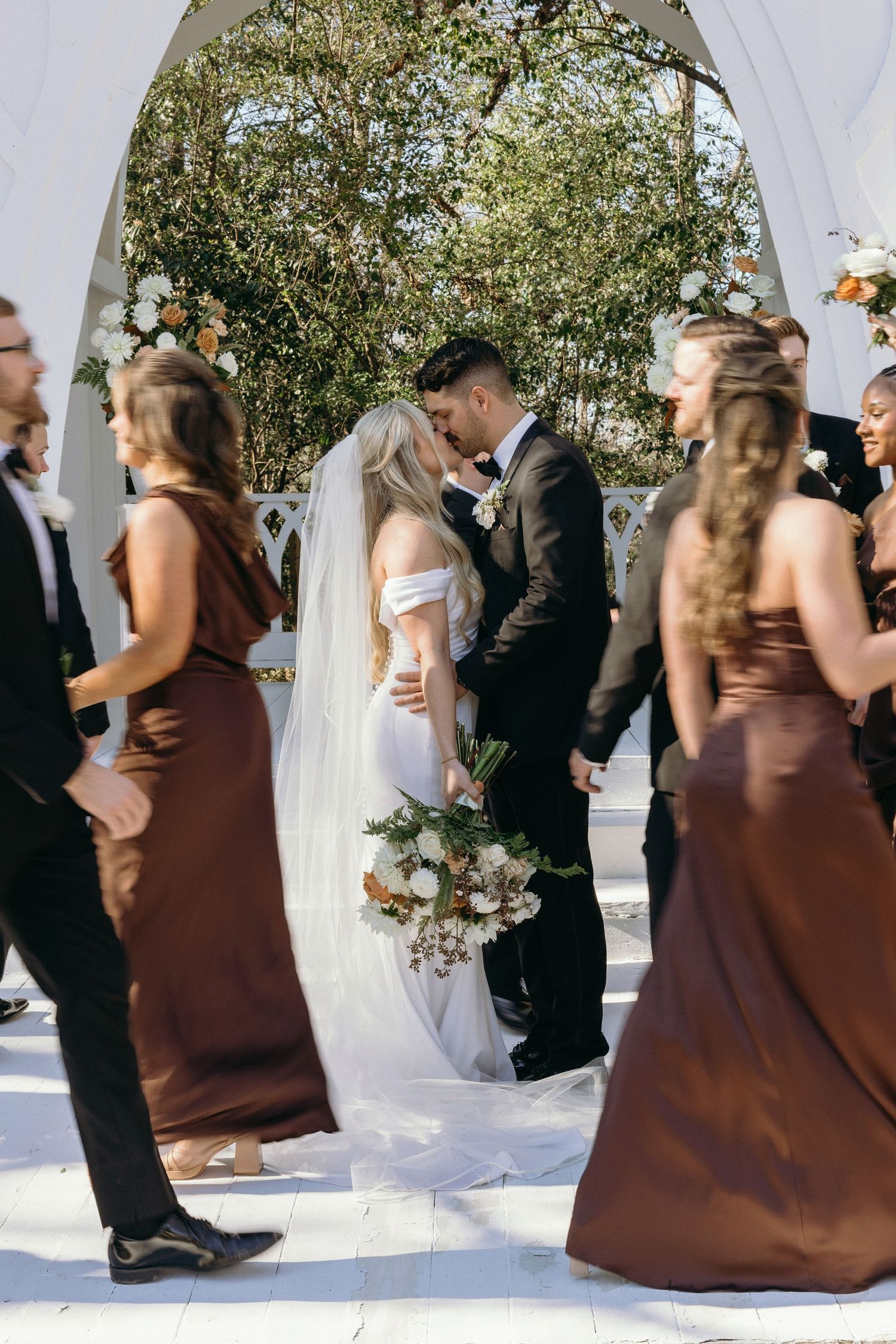 Bride and groom share first kiss at outdoor wedding altar, surrounded by bridesmaids in brown dresses at the Wildflower 301.