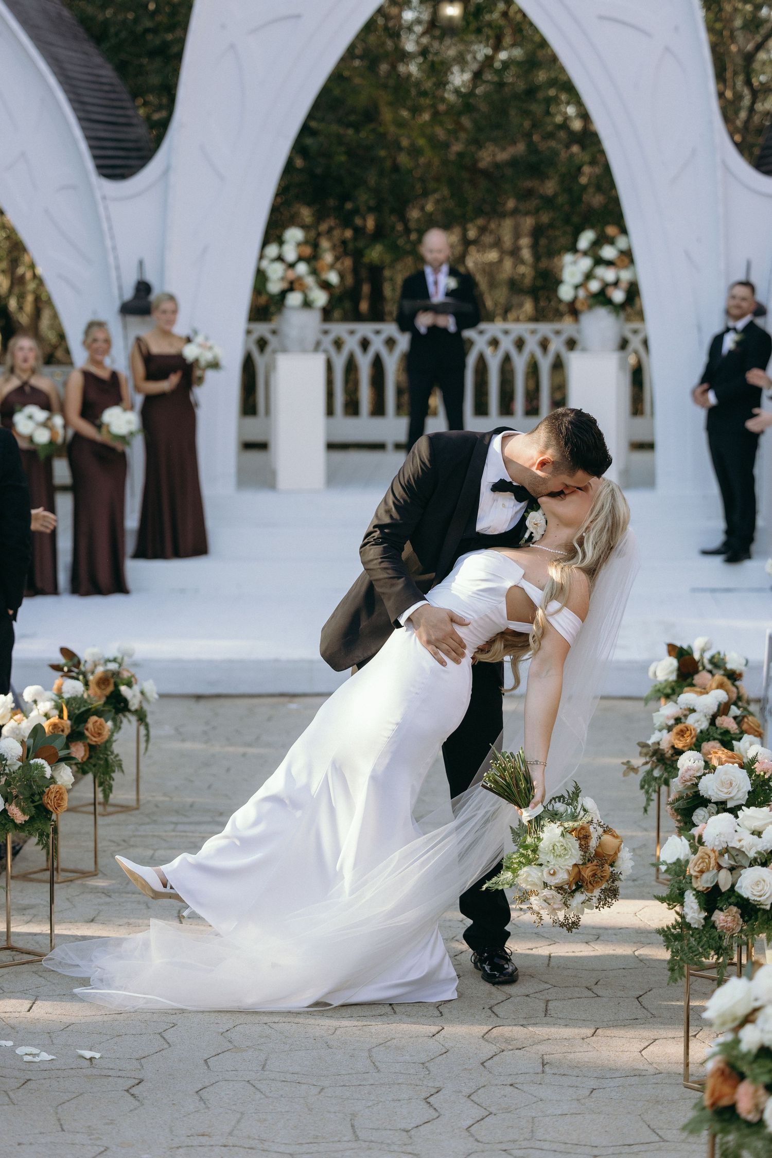 Groom dips bride for a kiss at outdoor wedding ceremony with white arch, floral arrangements, and wedding party in background.