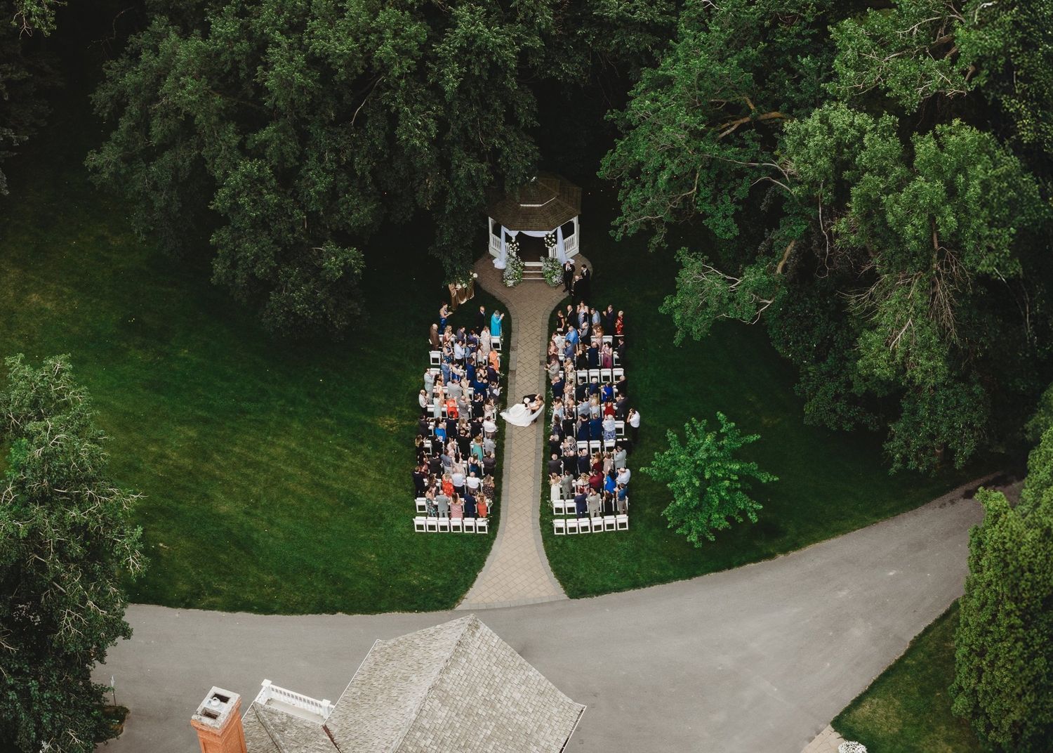 Aerial view of an outdoor wedding ceremony with guests seated in rows on a green lawn surrounded by lush trees.