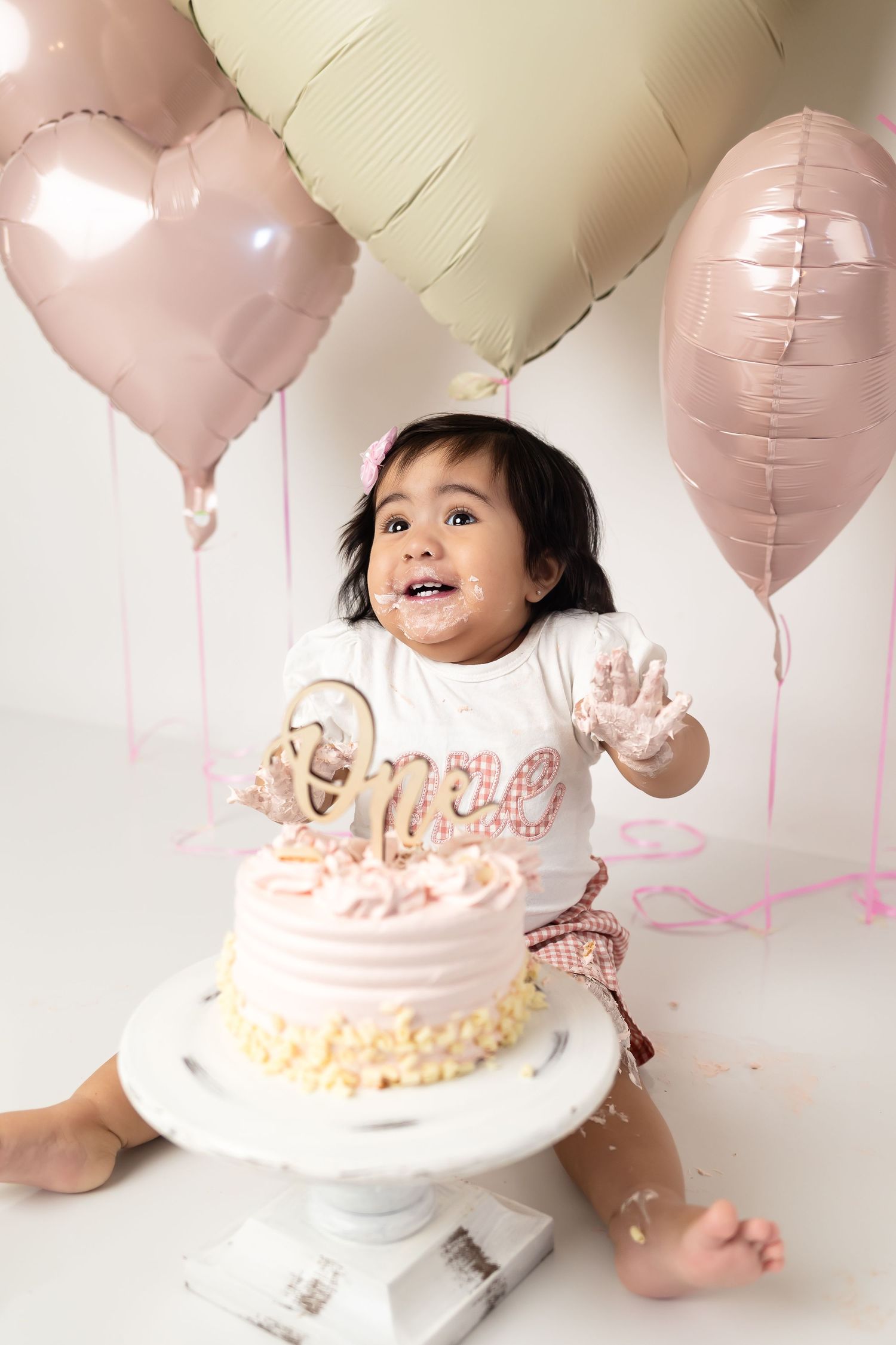 Baby girl celebrates first birthday smashing pink cake surrounded by heart balloons in white studio.