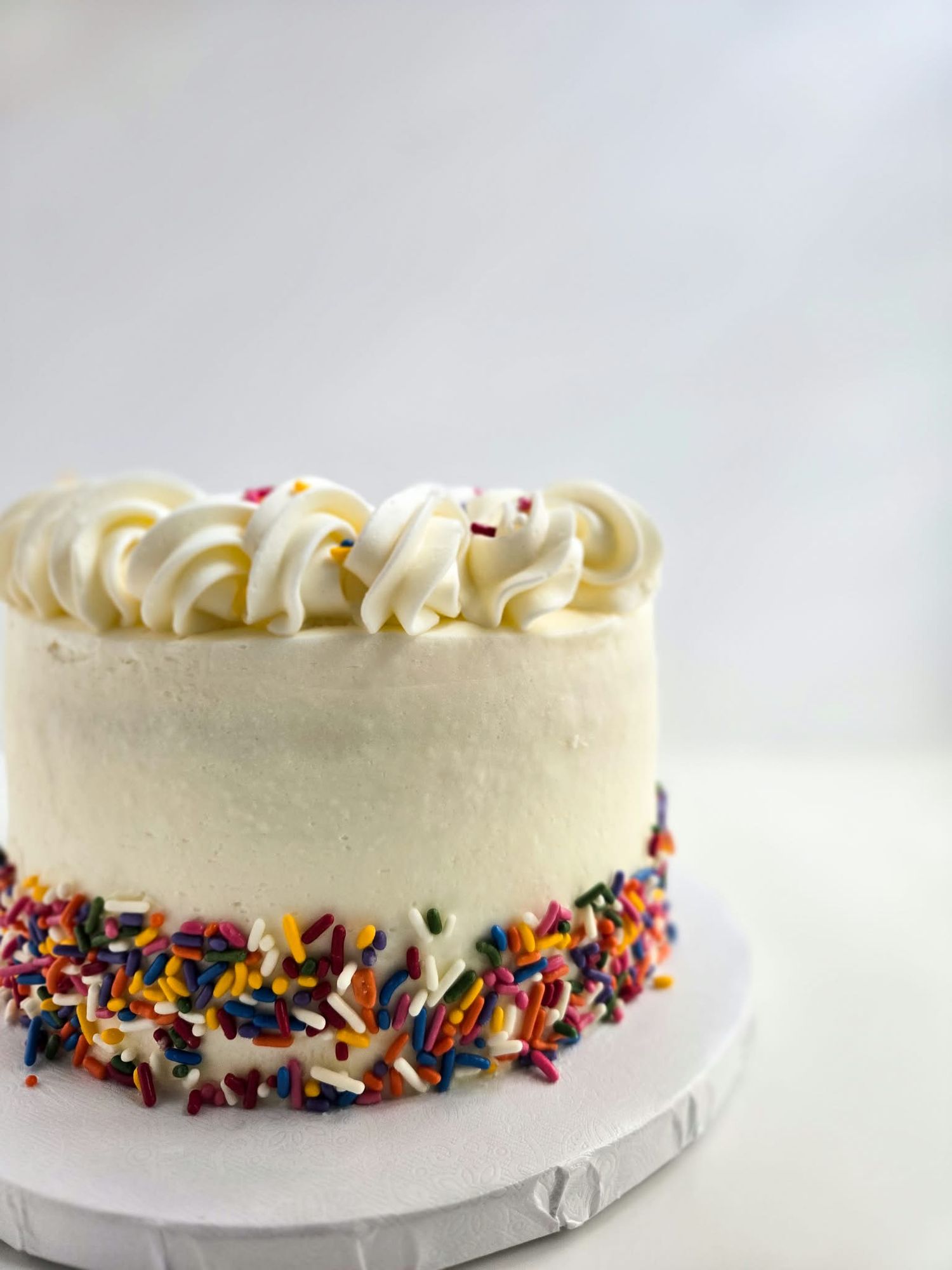 White frosted birthday cake with colorful rainbow sprinkles on base and swirled buttercream rosettes on top.
